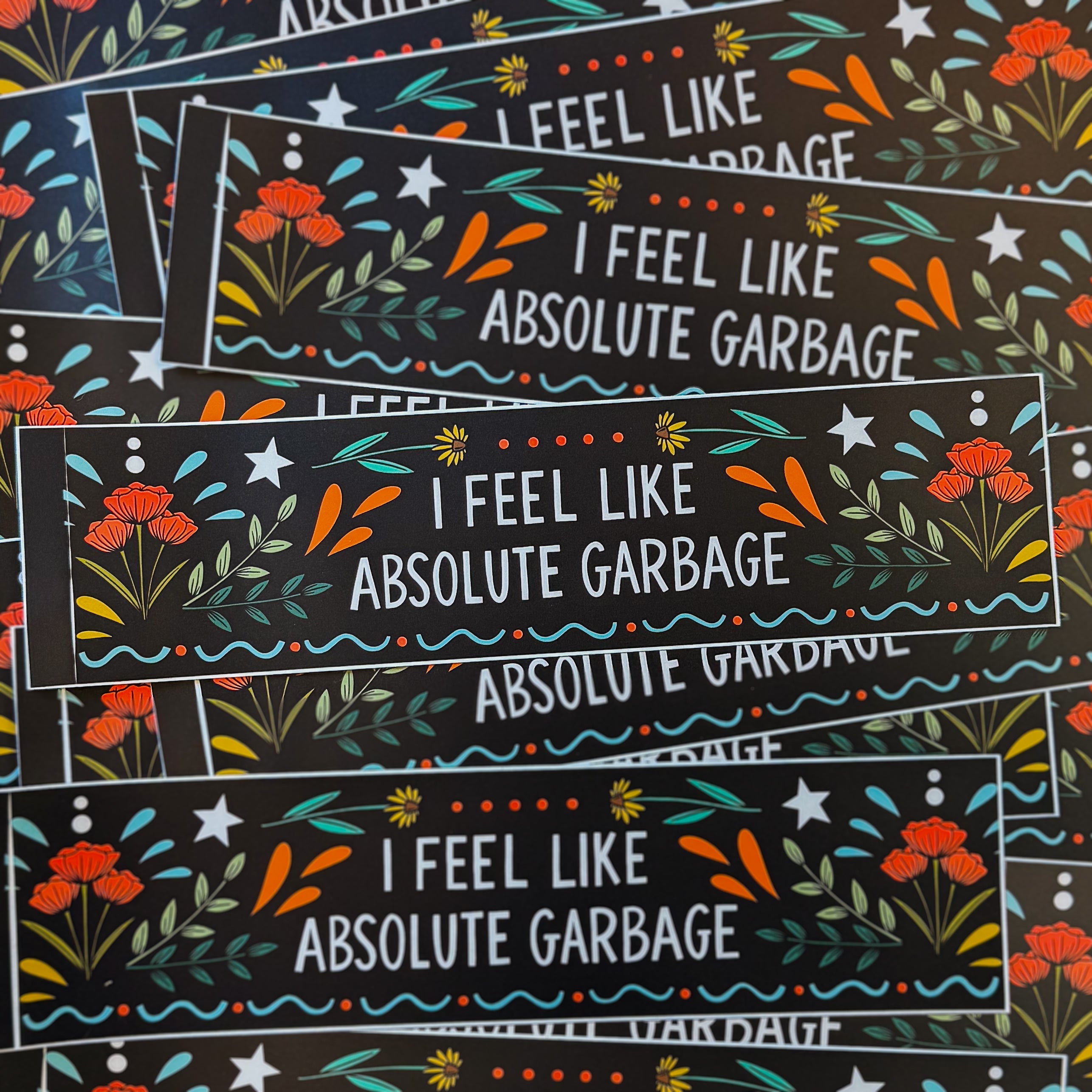 A black bumper sticker that says 'I FEEL LIKE ABSOLUTE GARBAGE' surrounded by flowers, stars, and flourishes.
