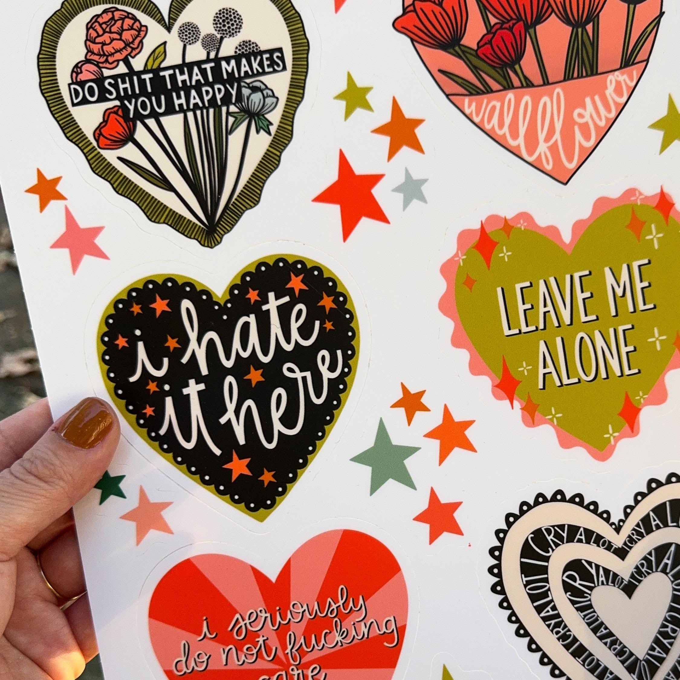 A hand holding a sticker sheet of six big hearts containing different silly sentiments with smaller star stickers in between.