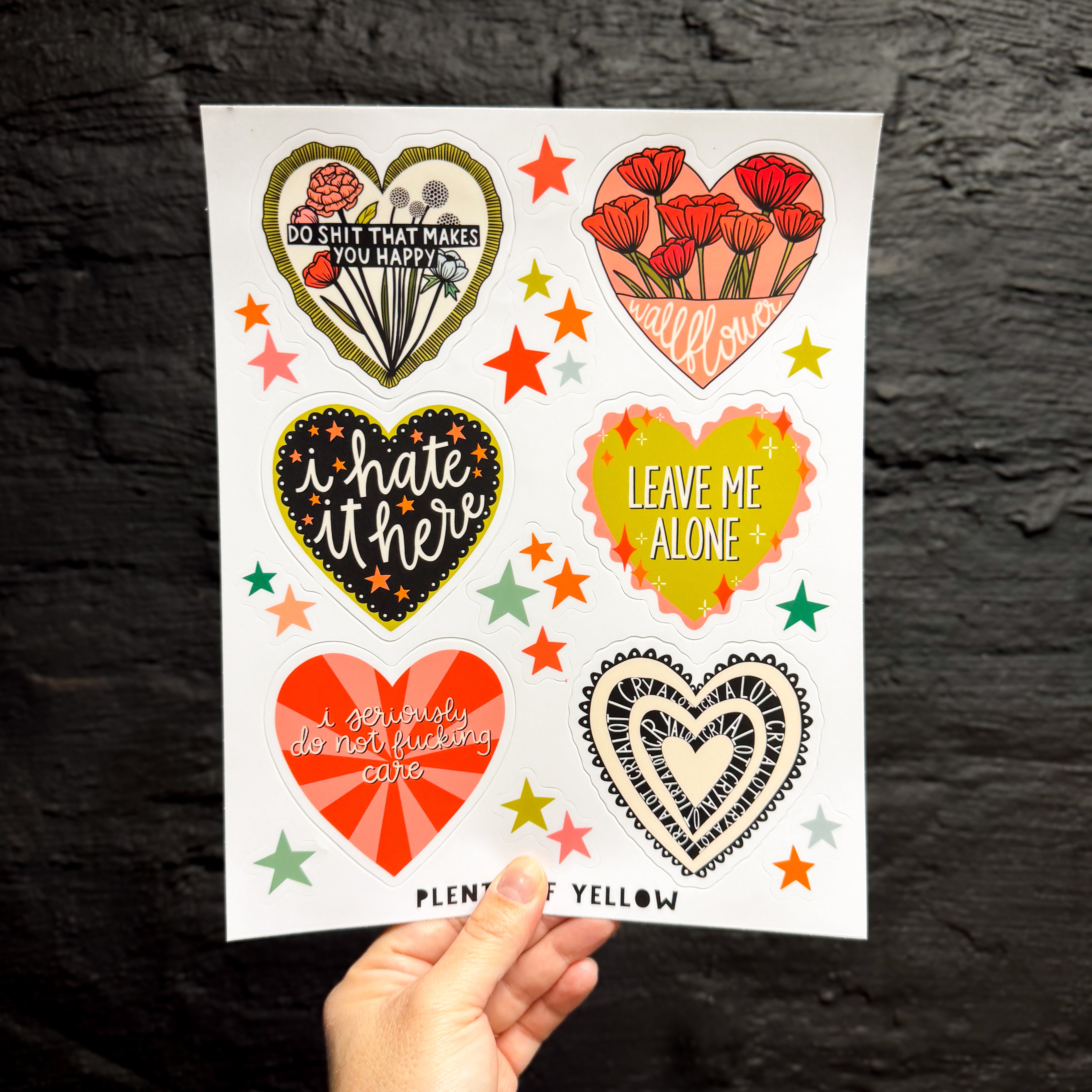 A hand holding a sticker sheet of six big hearts containing different silly sentiments with smaller star stickers in between.