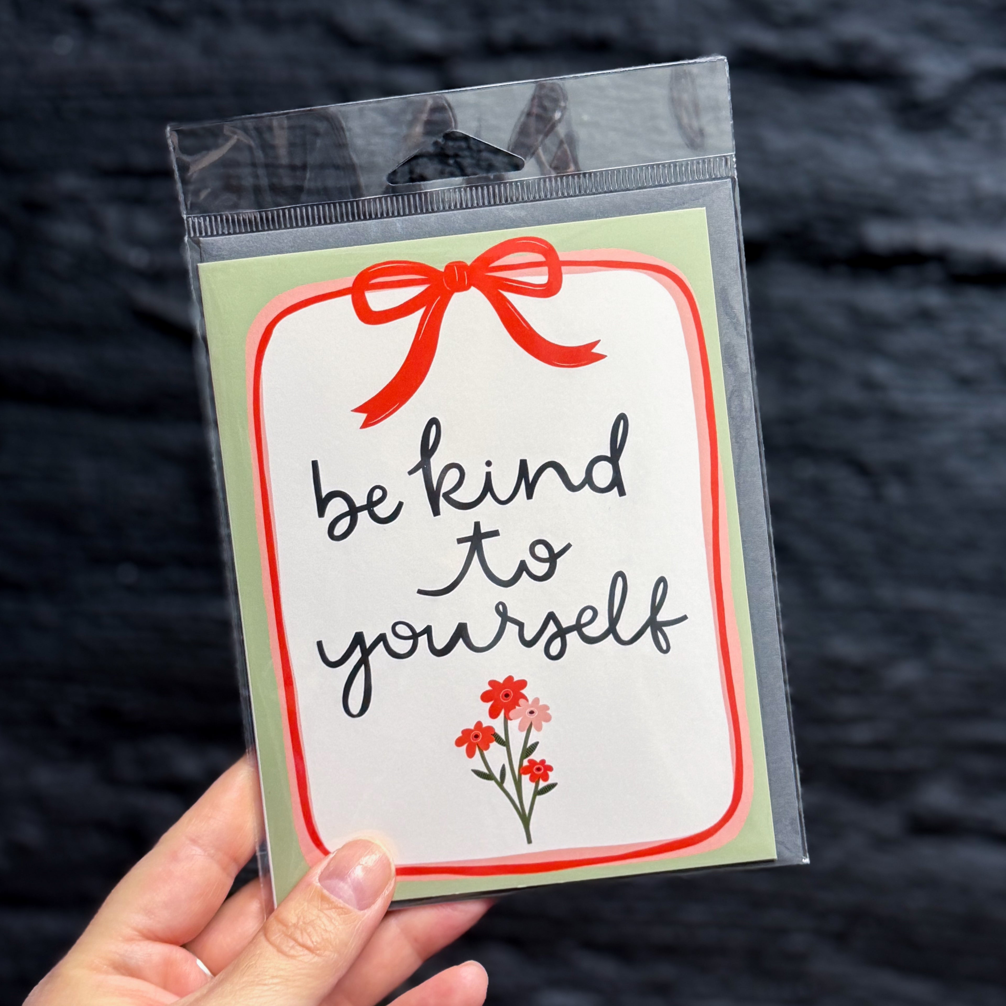 A hand holding a packaged greeting card with the phrase 'be kind to yourself' in black script on a white background, framed by a red border, a red bow illustration at the top, and small red flowers at the bottom, with a sage green border.