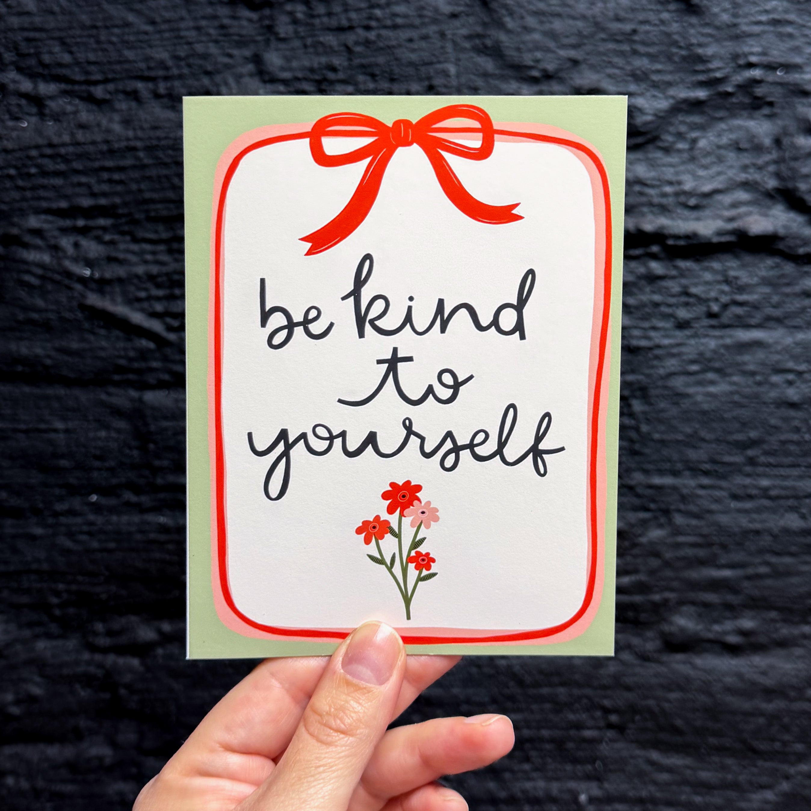 A hand holding a greeting card with the phrase 'be kind to yourself' in black script on a white background, framed by a red border, a red bow illustration at the top, and small red flowers at the bottom, with a sage green border.