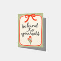 A mockup of a greeting card with the phrase 'be kind to yourself' in black script on a white background, framed by a red border, a red bow illustration at the top, and small red flowers at the bottom, with a sage green border.