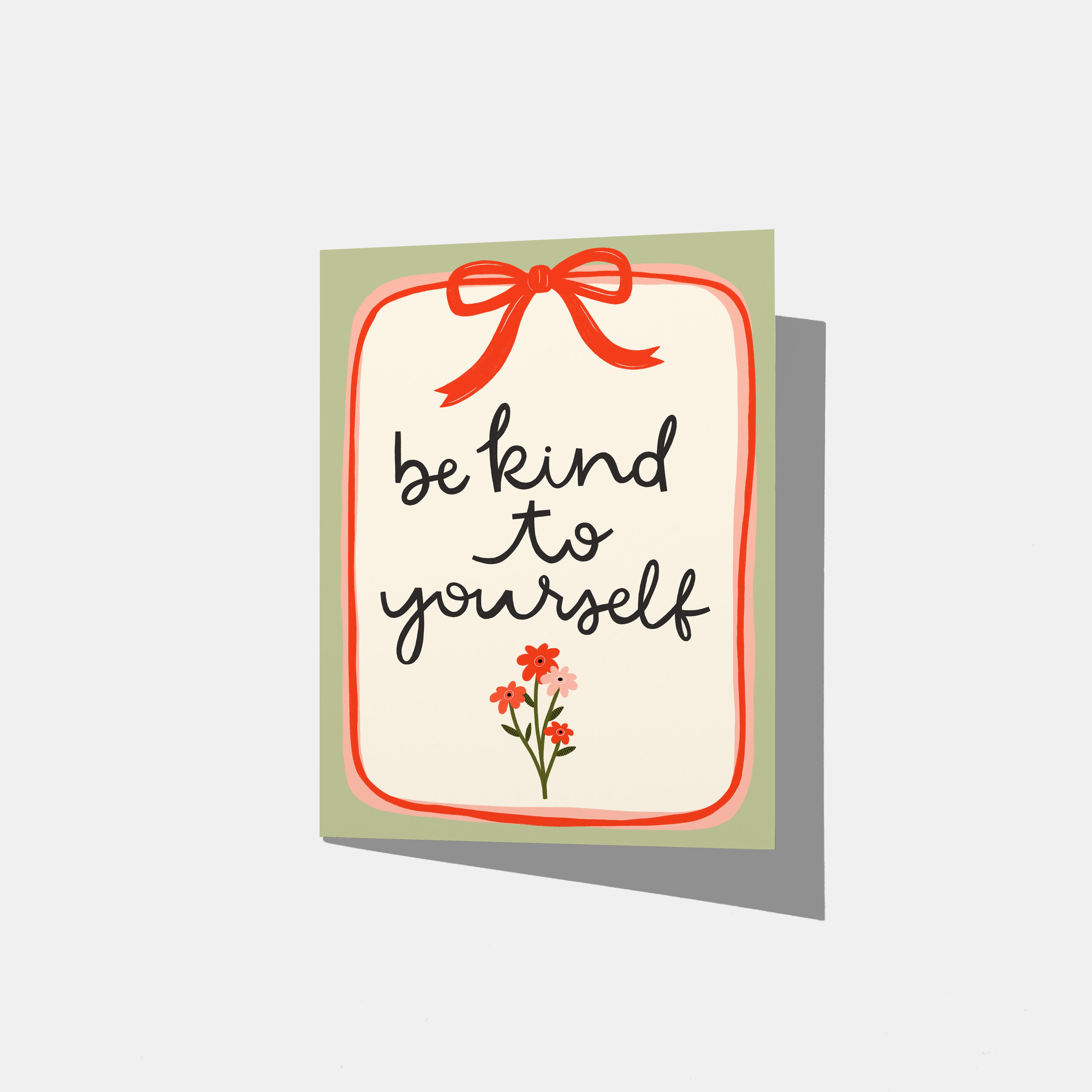 A mockup of a greeting card with the phrase 'be kind to yourself' in black script on a white background, framed by a red border, a red bow illustration at the top, and small red flowers at the bottom, with a sage green border.