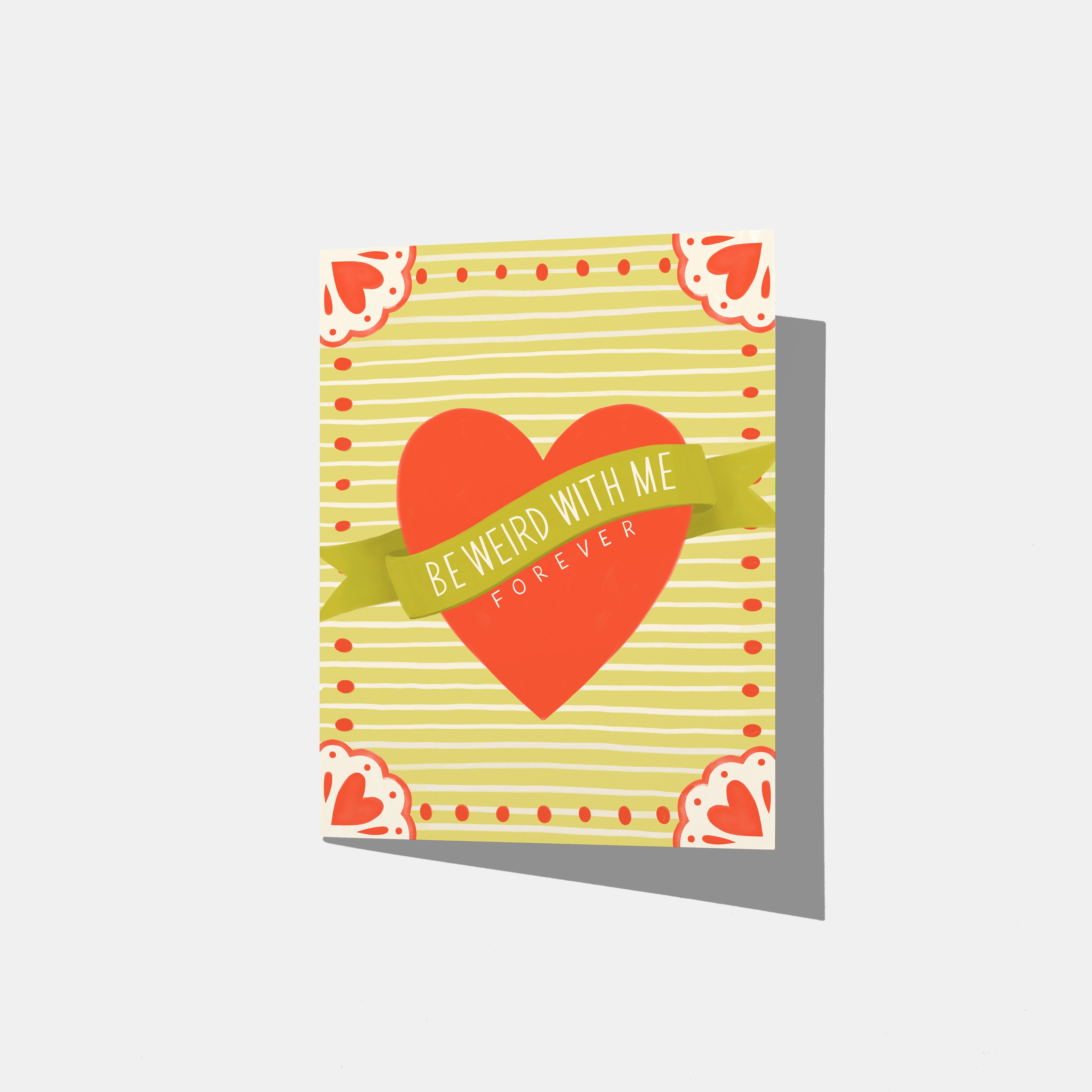 A mockup of a greeting card that says 'BE WEIRD WITH ME FOREVER' on a green banner across a red heart. The background is yellow/green strips with red hearts and embellishments in the corners.