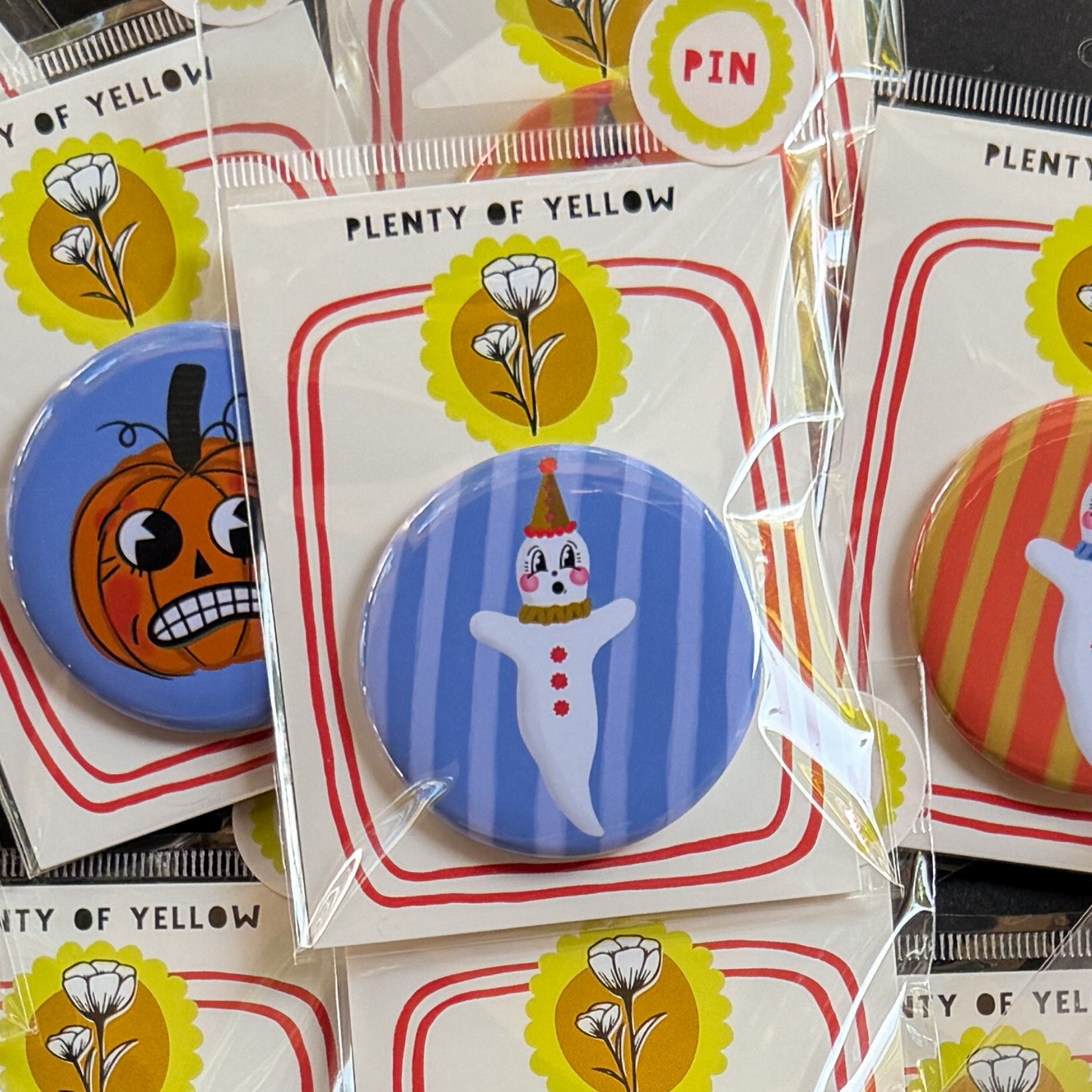 A round pinback button in clear plastic packaging, featuring a ghostie clown with a blue striped background. The pin is mounted on a white backing card with red borders and a yellow logo, under the brand name 'PLENTY OF YELLOW'.