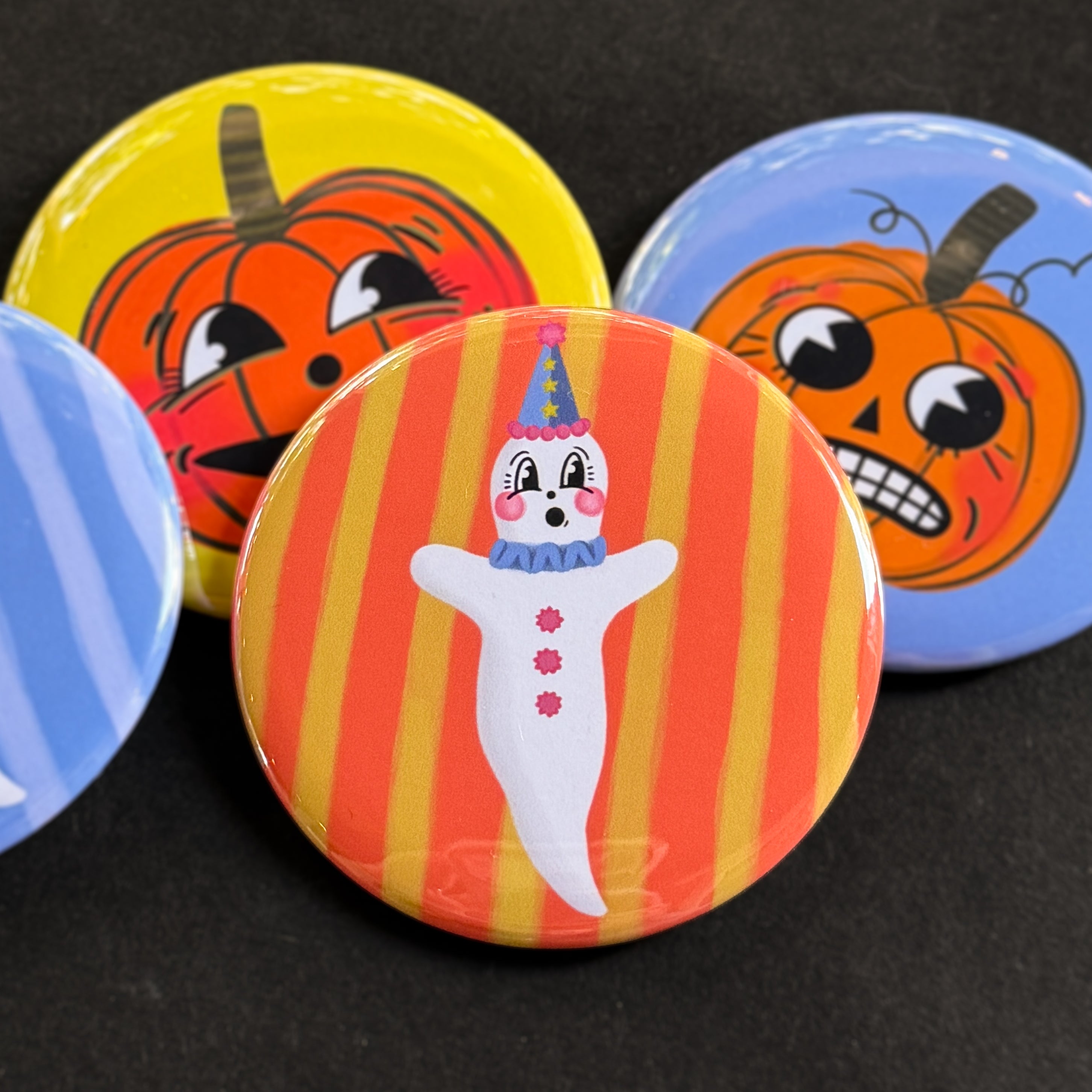 A round pinback button featuring a ghostie clown with an orange and yellow striped background. The pin is surrounded by other pin designs.