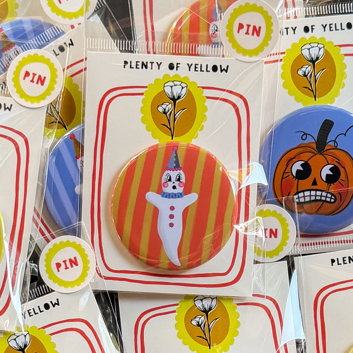 A round pinback button in clear plastic packaging, featuring a ghostie clown with an orange and yellow striped background. The pin is mounted on a white backing card with red borders and a yellow logo, under the brand name 'PLENTY OF YELLOW'.