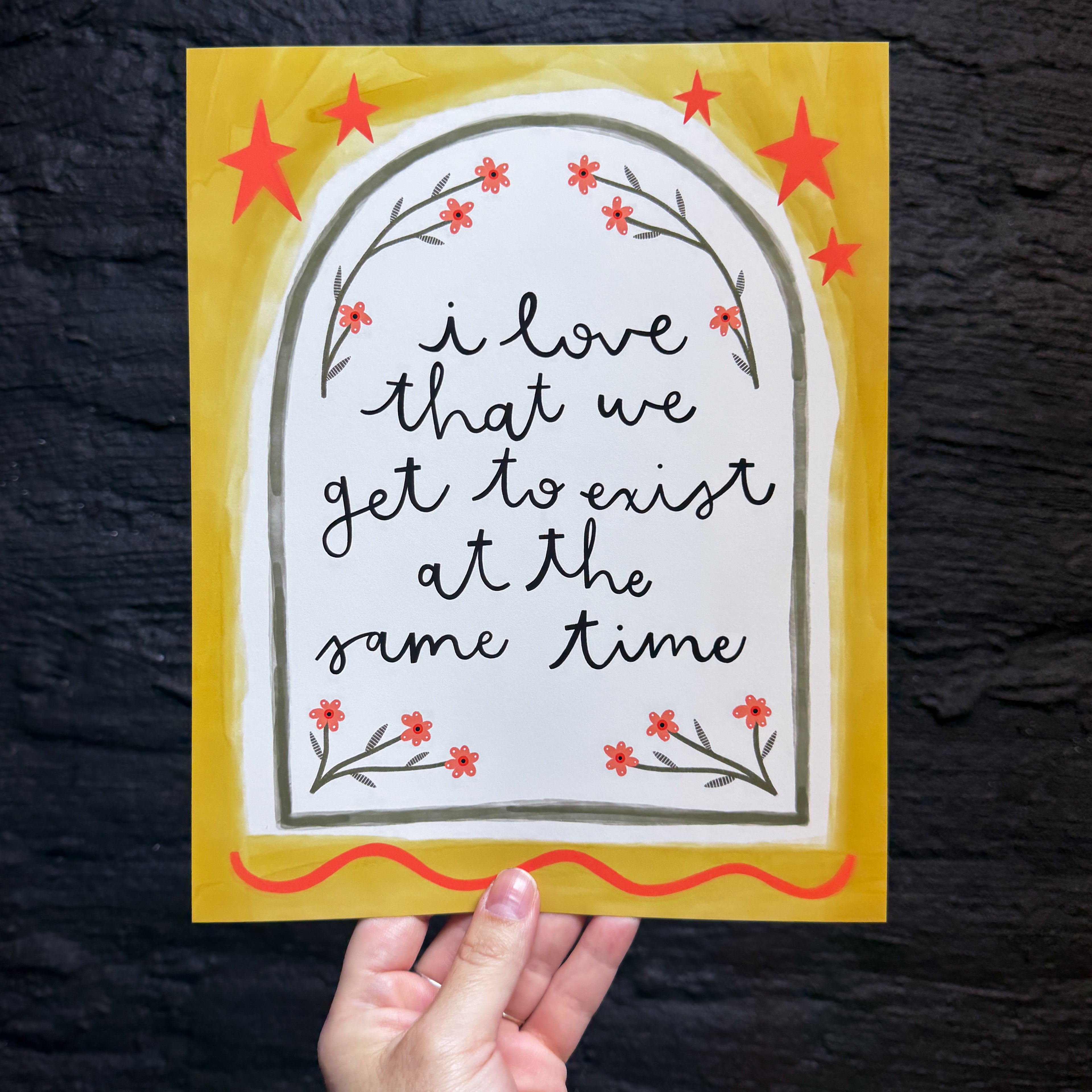A hand holding an 8x10 yellow print that says 'i love that we get to exist at the same time' inside a cream and green arch with little flowers framing it and red stars and embellishments on the edge.