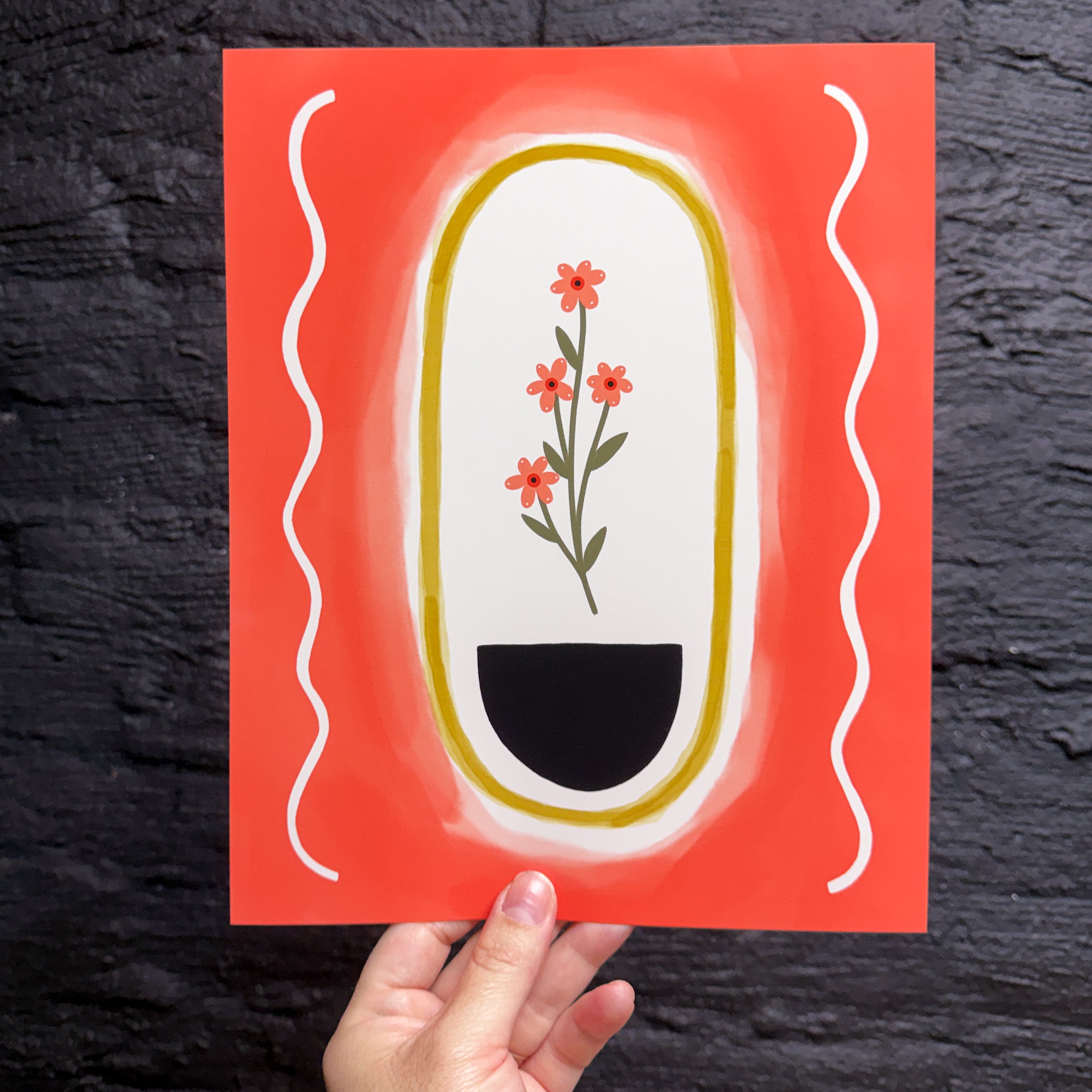 A hand holding an 8x10 art print with a red/orange background, cream oval and squiggles on the sides. Inside the oval is a yellow outline and four pink flowers sitting above a black half circle. 