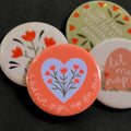 A round pinback button featuring a coral background with a blue heart in the middle behind a bunch of red flowers. The text ‘i did not sign up for this.’ sits on the bottom edge. The pin is surrounded by other pin designs.