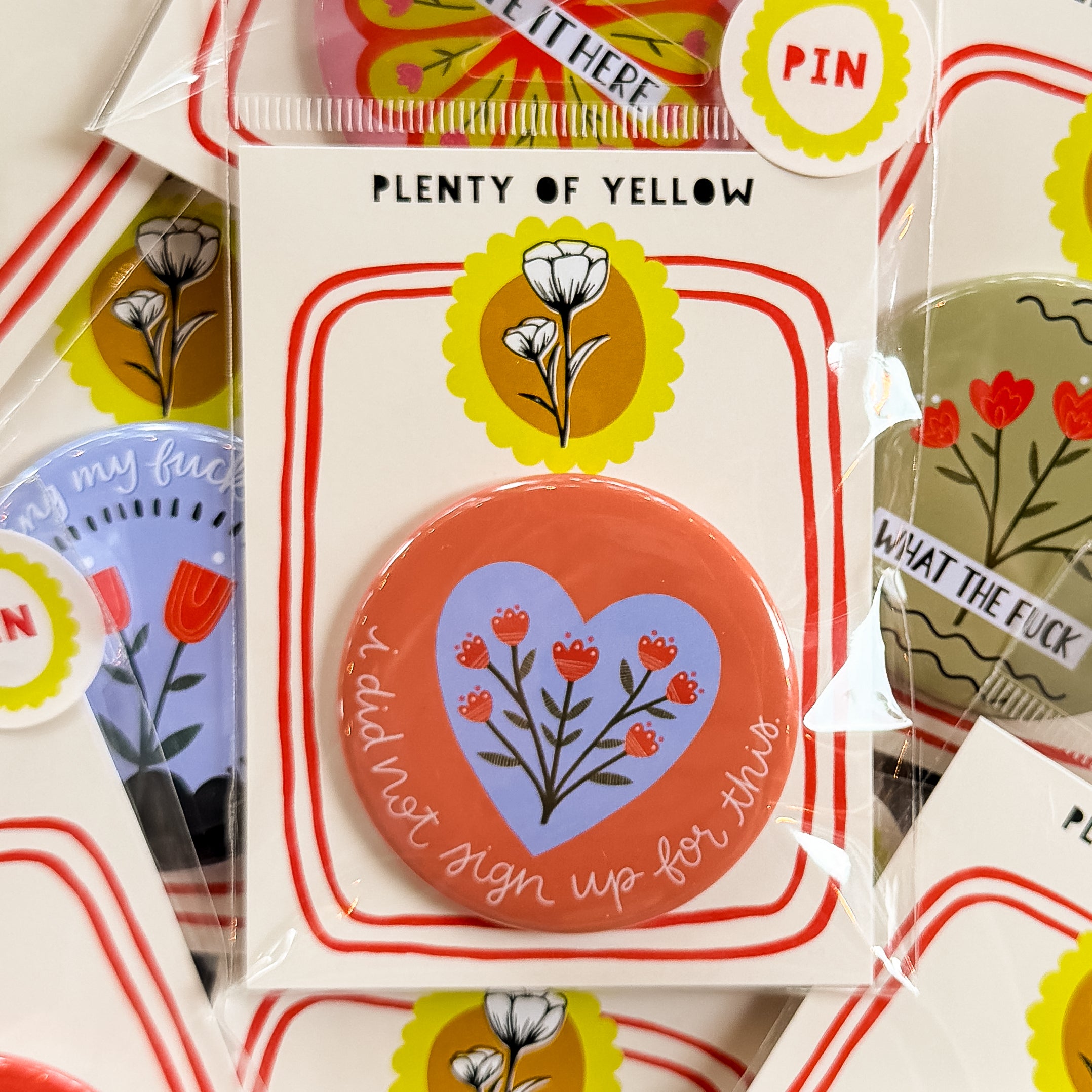 A round pinback button in clear plastic packaging, featuring a a coral background with a blue heart in the middle behind a bunch of red flowers. The text ‘i did not sign up for this.’ sits on the bottom edge. The pin is mounted on a white backing card with red borders and a yellow logo, under the brand name 'PLENTY OF YELLOW'.