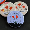 A round pinback button featuring a cornflower blue background with red flowers surrounded by a black dashed circle with a black scalloped bottom. The text ‘doing my fucking best’ sits across the top. The pin is surrounded by other pin designs.