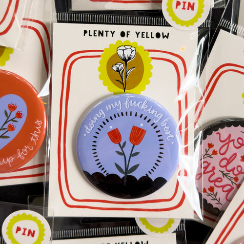 A round pinback button in clear plastic packaging, featuring a cornflower blue background with red flowers surrounded by a black dashed circle with a black scalloped bottom. The text ‘doing my fucking best’ sits across the top. The pin is mounted on a white backing card with red borders and a yellow logo, under the brand name 'PLENTY OF YELLOW'.