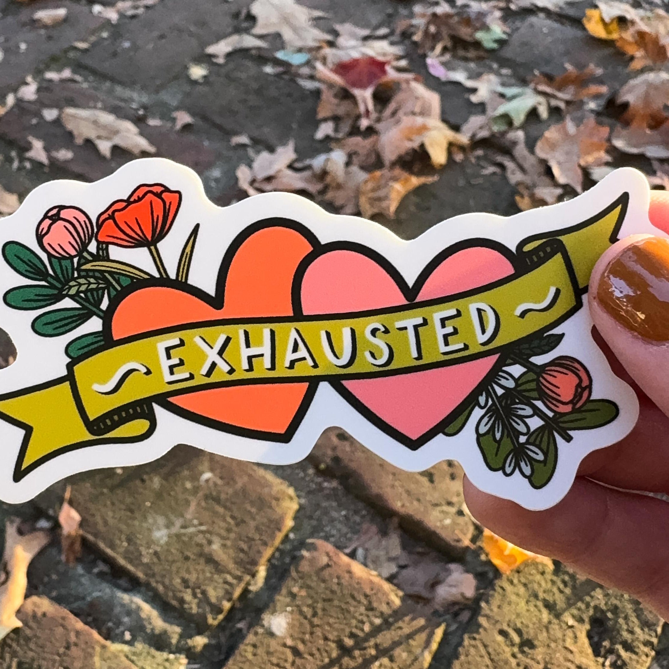 A hand holding a sticker with two hearts and flowers with 'exhausted' written across it.