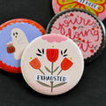 A round pinback button featuring a cream background with red flowers with yellow and pink embellishments. The text ‘EXHAUSTED’ sits across it. The pin is surrounded by other pin designs.
