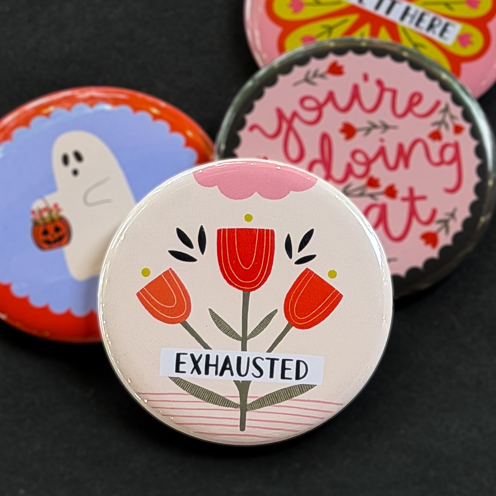 A round pinback button featuring a cream background with red flowers with yellow and pink embellishments. The text ‘EXHAUSTED’ sits across it. The pin is surrounded by other pin designs.
