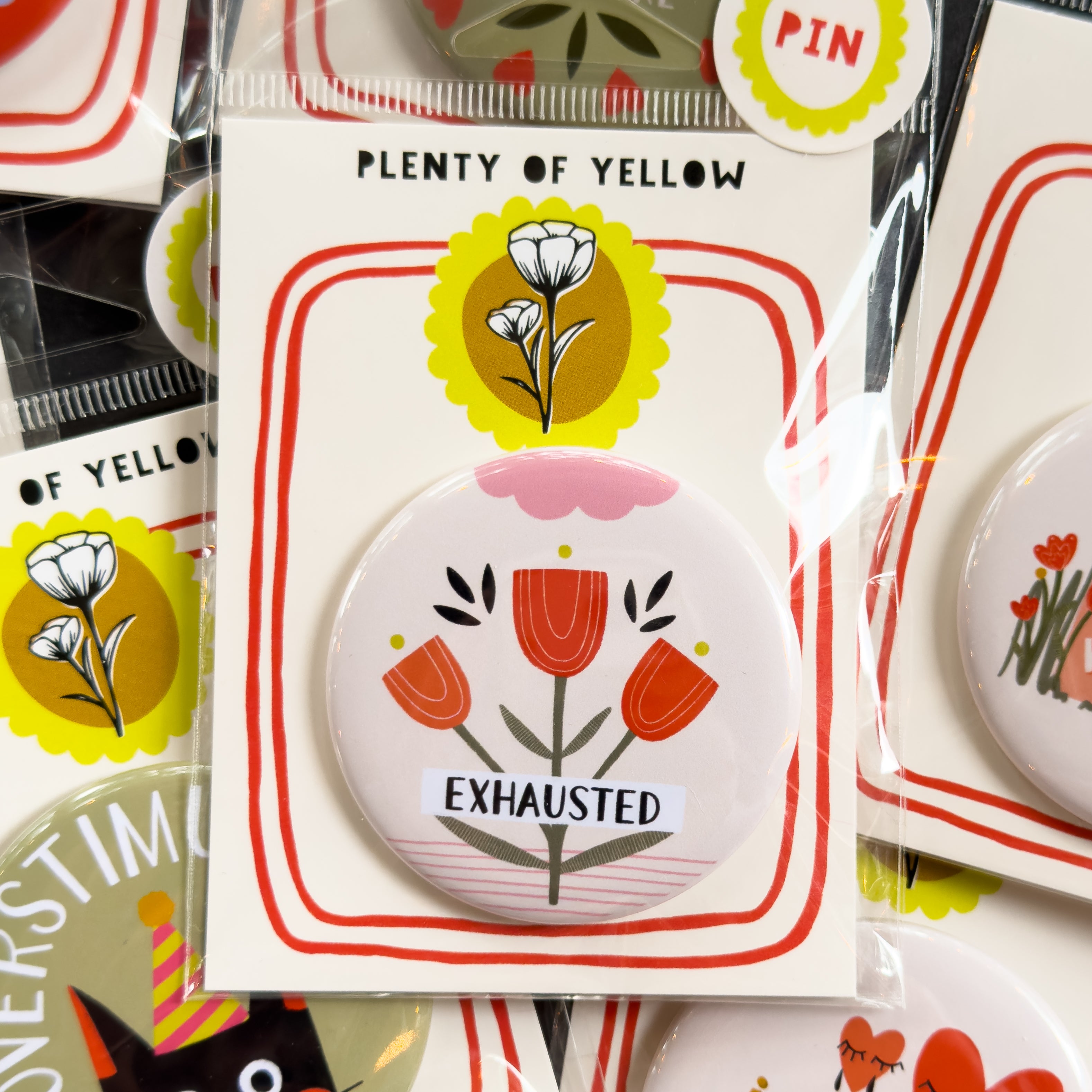 A round pinback button in clear plastic packaging, featuring a cream background with red flowers with yellow and pink embellishments. The text ‘EXHAUSTED’ sits across it. The pin is mounted on a white backing card with red borders and a yellow logo, under the brand name 'PLENTY OF YELLOW'.