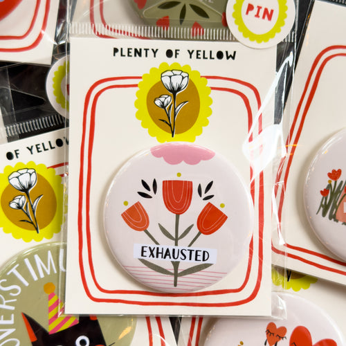A round pinback button in clear plastic packaging, featuring a cream background with red flowers with yellow and pink embellishments. The text ‘EXHAUSTED’ sits across it. The pin is mounted on a white backing card with red borders and a yellow logo, under the brand name 'PLENTY OF YELLOW'.