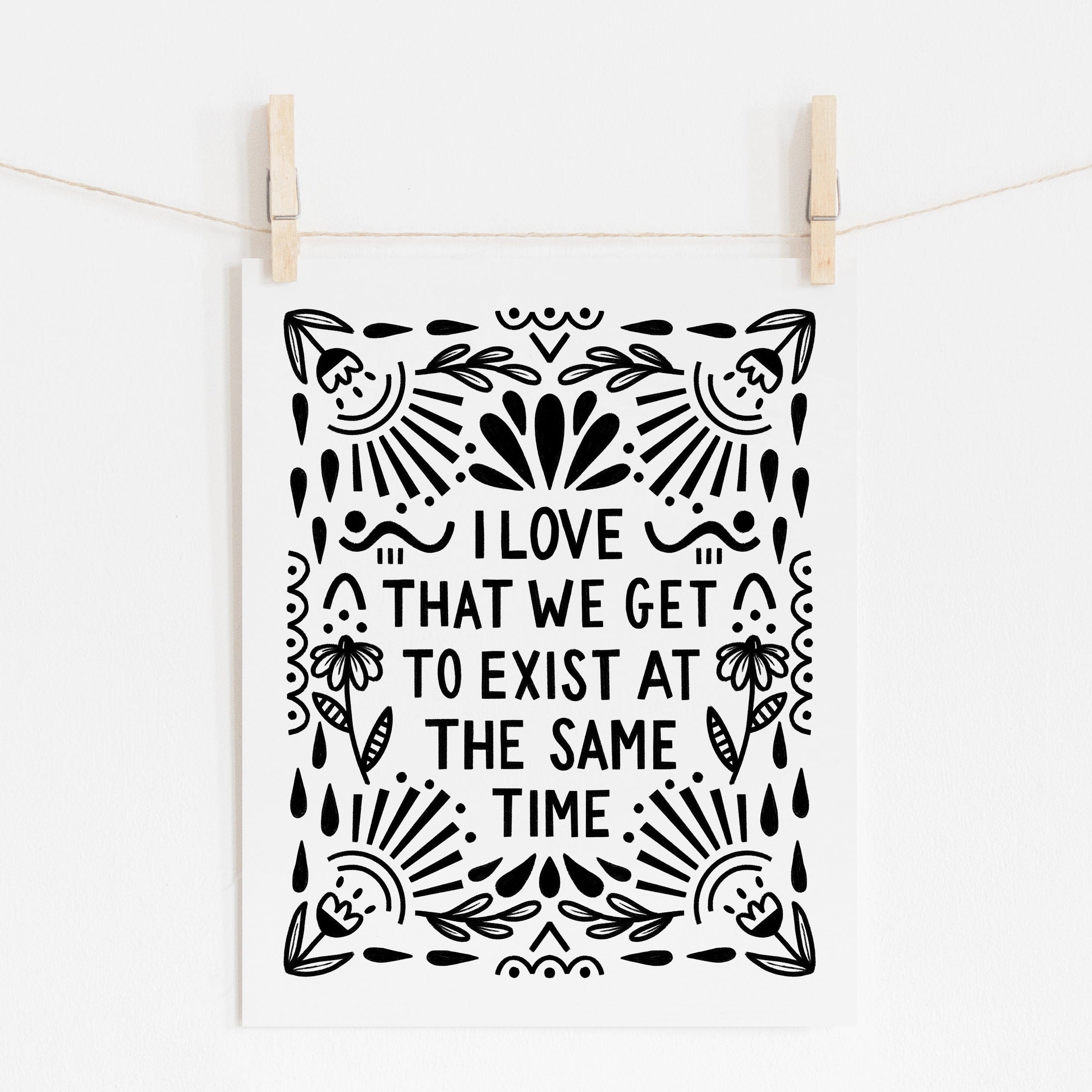 A mockup of an 8x10 art print that says 'I LOVE THAT WE GET TO EXIST AT THE SAME TIME' in black on white paper with floral and whimsy embellishments around it.