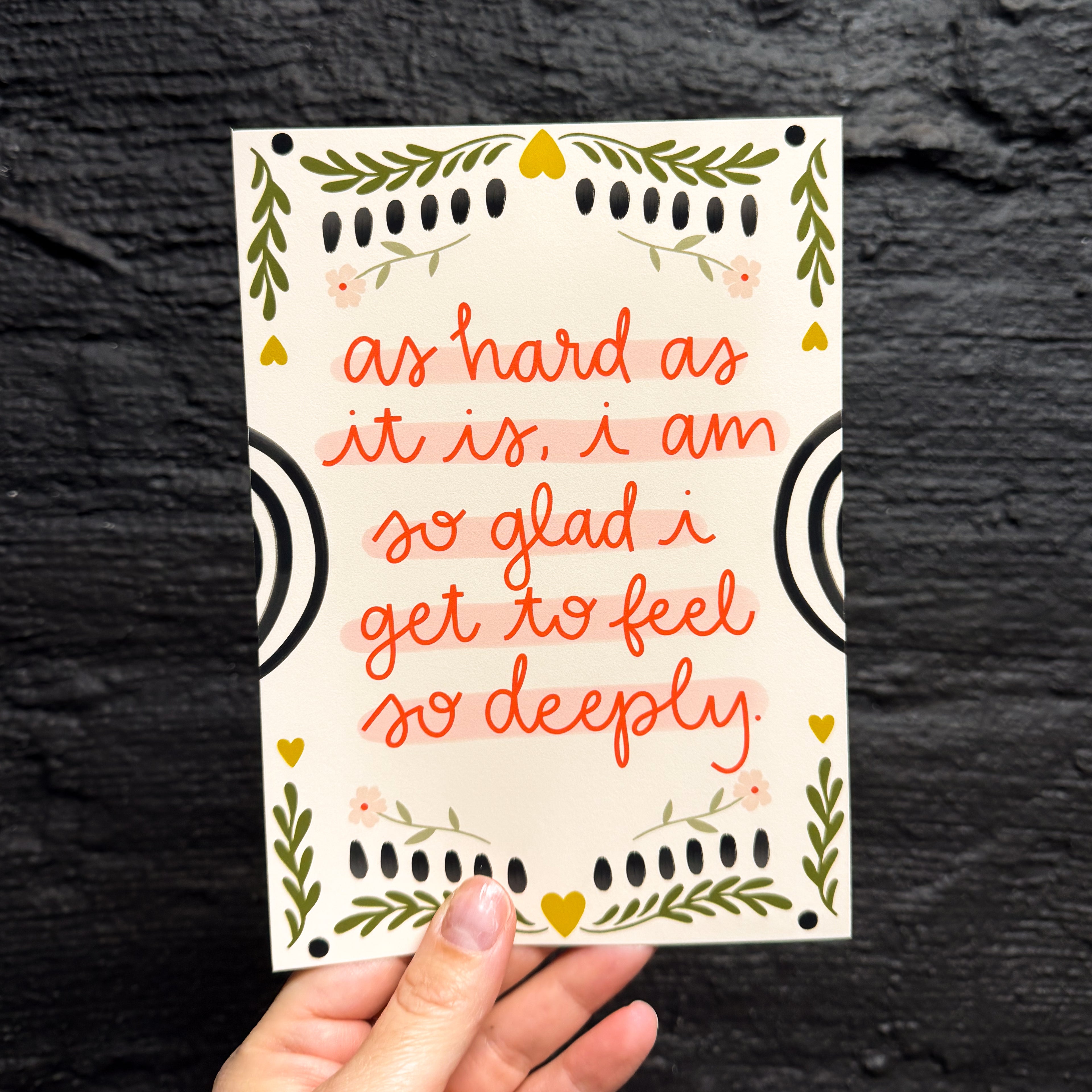 A hand holding a 5x7 art print with a cream background. It says 'as hard as it is, i am so glad i get to feel so deeply.' in red with embellishments of flowers and leaves as a border.