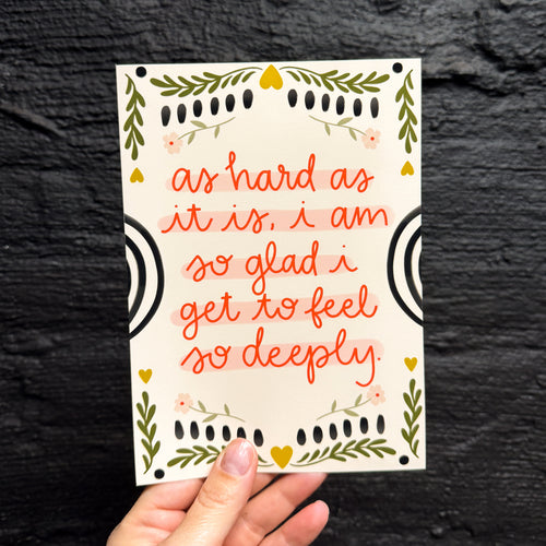 A hand holding a 5x7 art print with a cream background. It says 'as hard as it is, i am so glad i get to feel so deeply.' in red with embellishments of flowers and leaves as a border.