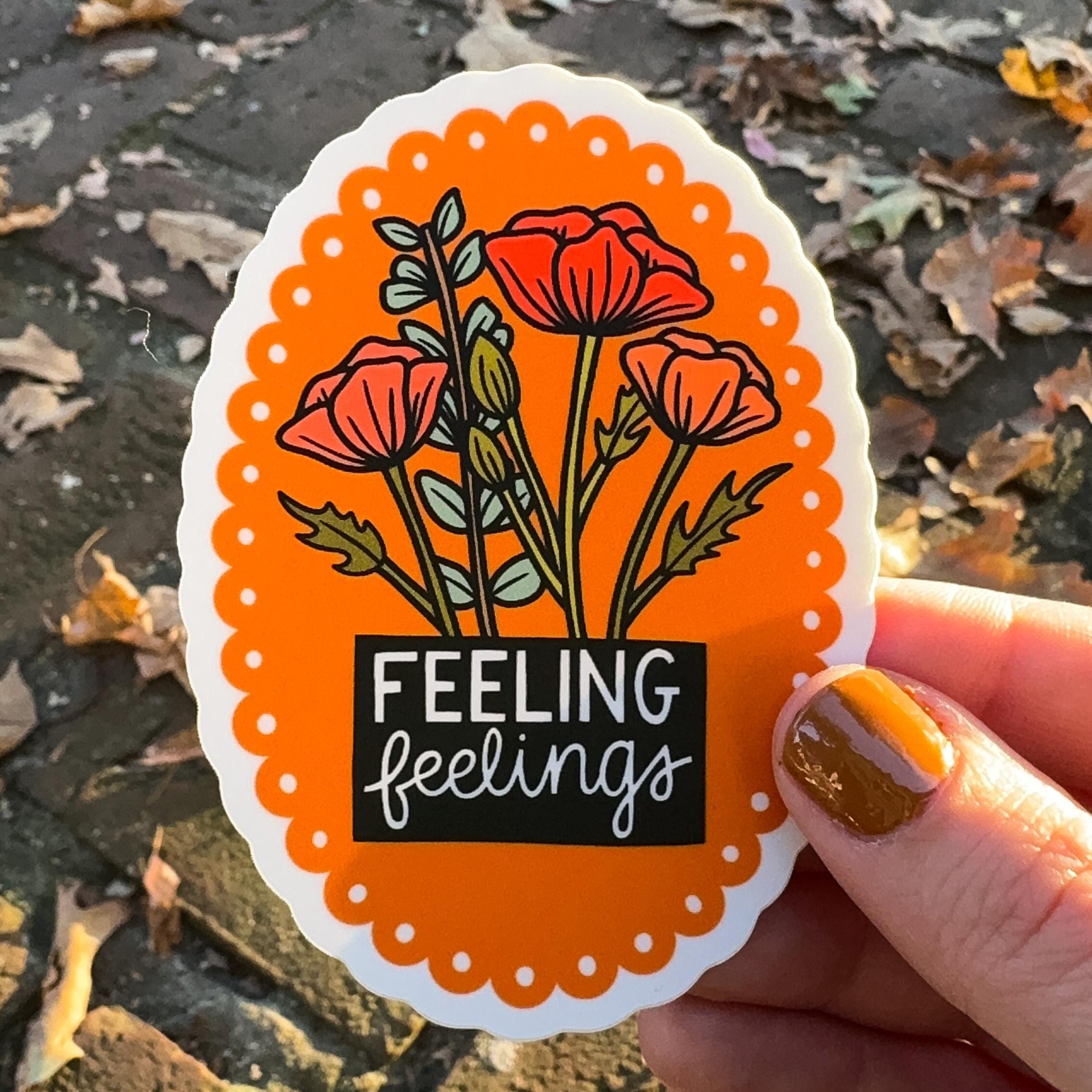 A hand holding a sticker with scalloped floral design and 'FEELING feelings' text on an orange background.