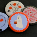 A round pinback button featuring a red background with a blue scalloped circle in the middle behind a ghost with a trick-or-treat pumpkin filled with flowers. The pin is surrounded by other pin designs.