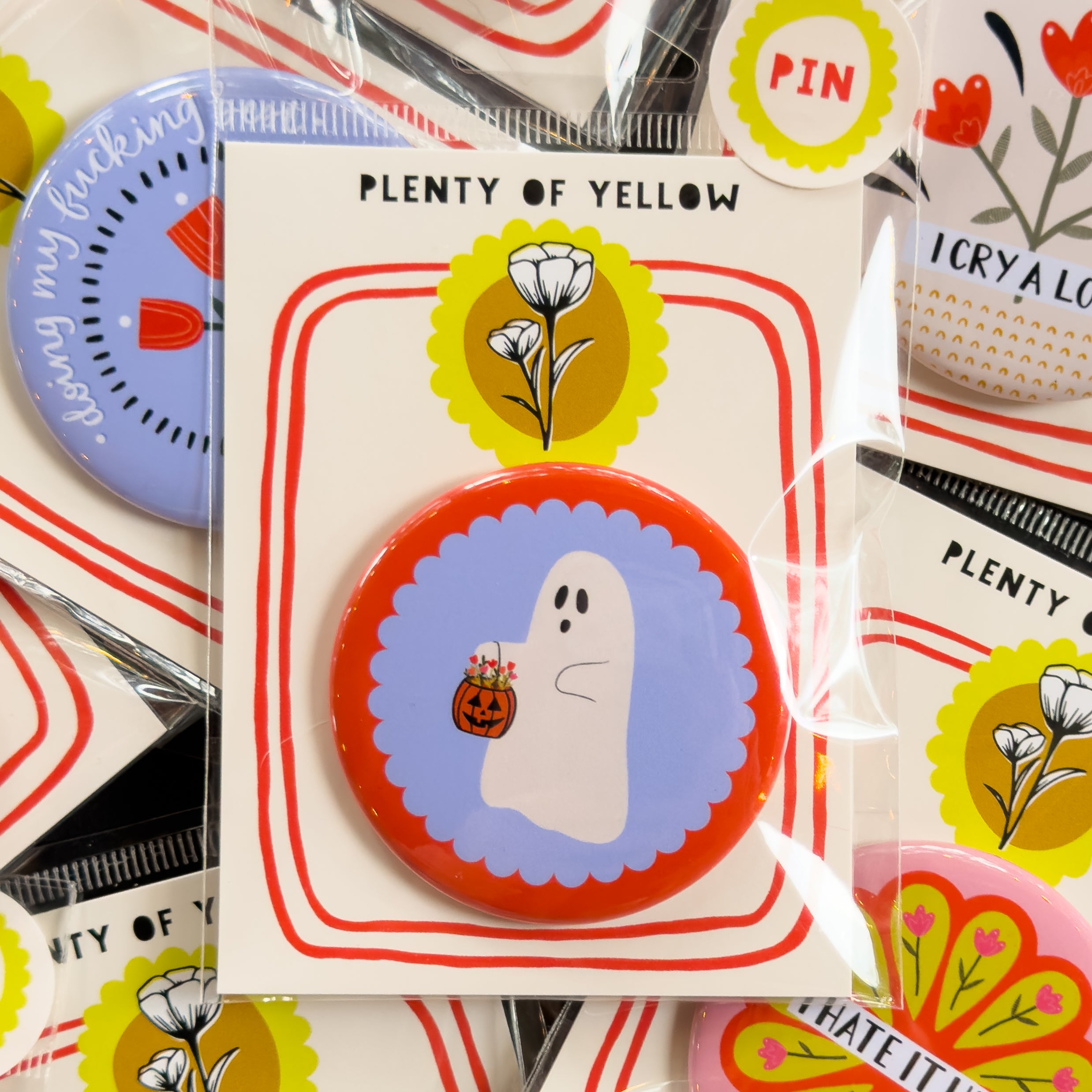 A round pinback button in clear plastic packaging, featuring a red background with a blue scalloped circle in the middle behind a ghost with a trick-or-treat pumpkin filled with flowers. The pin is mounted on a white backing card with red borders and a yellow logo, under the brand name 'PLENTY OF YELLOW'.