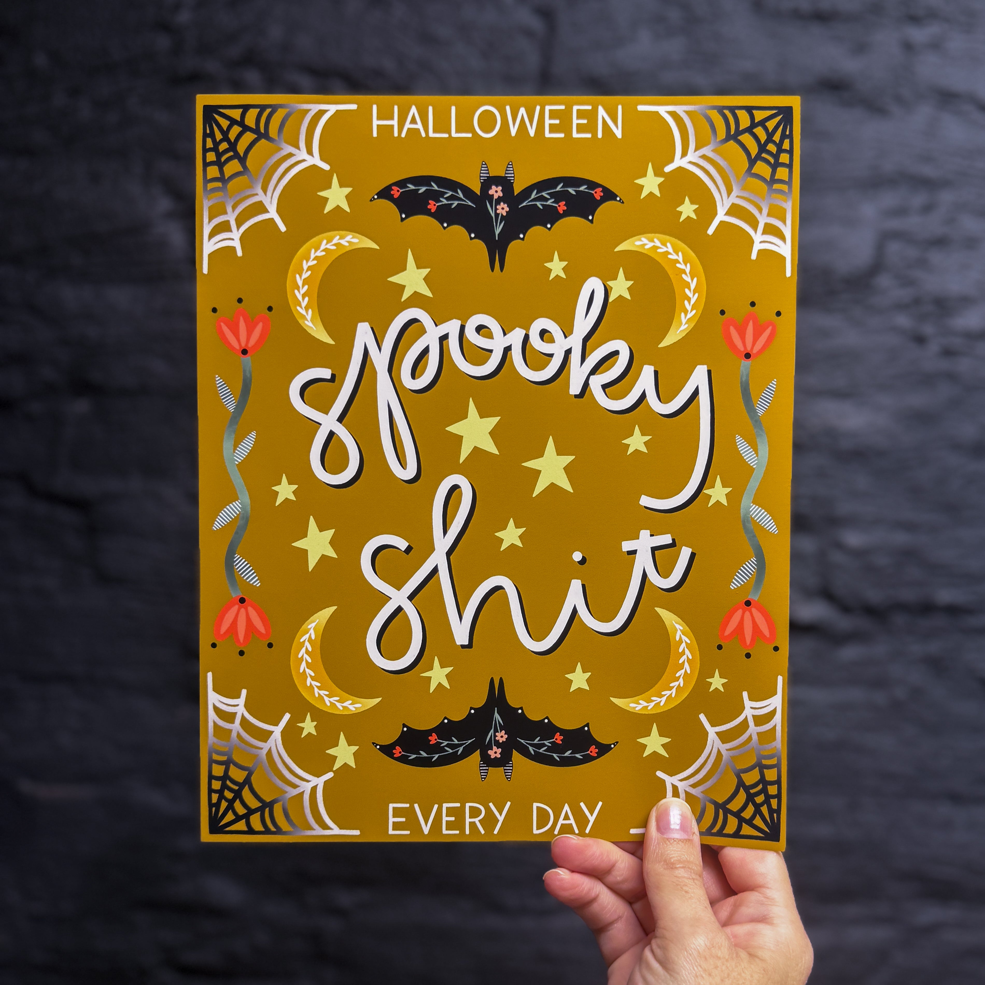 A hand holding an 8x10 Halloween art print with a yellow background that says "spooky shit" in the middle and "HALLOWEEN EVERY DAY" framing the top and bottom. There are spiderwebs, bats, crescent moons, stars, and flowers embellishing it. 