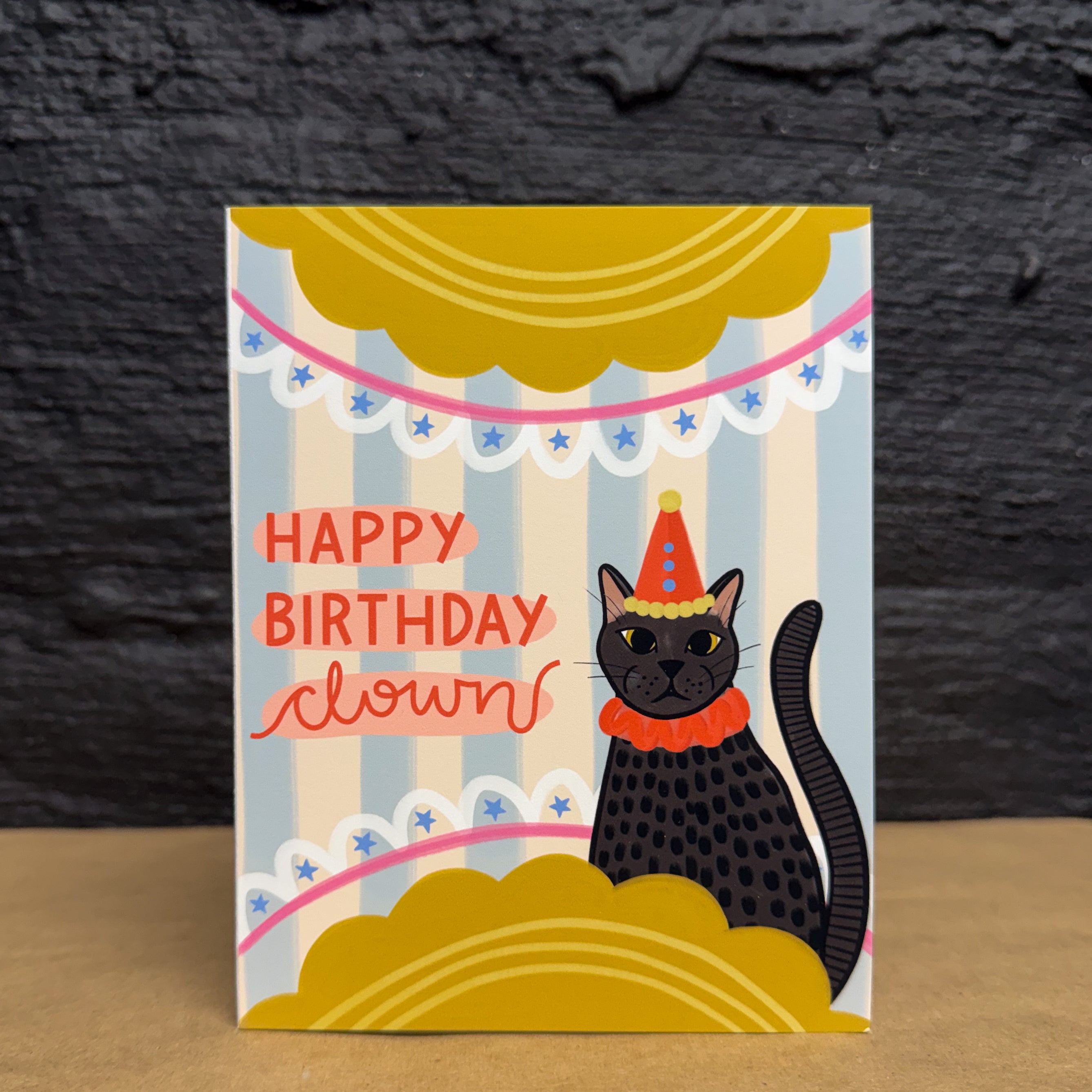 A birthday card with a black cat wearing a party hat and 'Happy Birthday Clown' text on a blue striped background with yellow scalloped half circles on the top and bottom with stars and line embellishments.