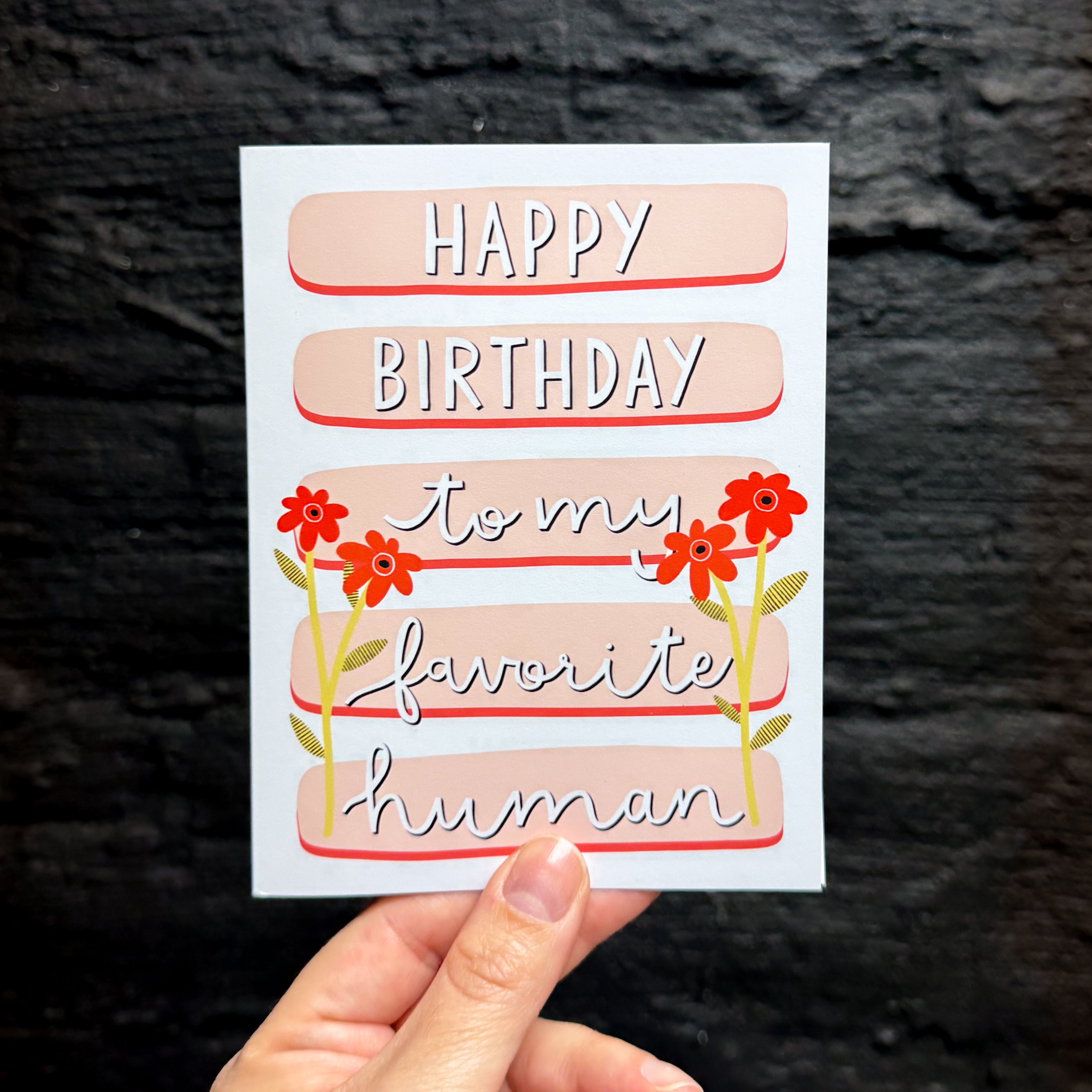 A hand holding a birthday card with the text 'HAPPY BIRTHDAY to my favorite human' in stylized lettering, on a background of peach-colored horizontal bards, decorated with red flowers.