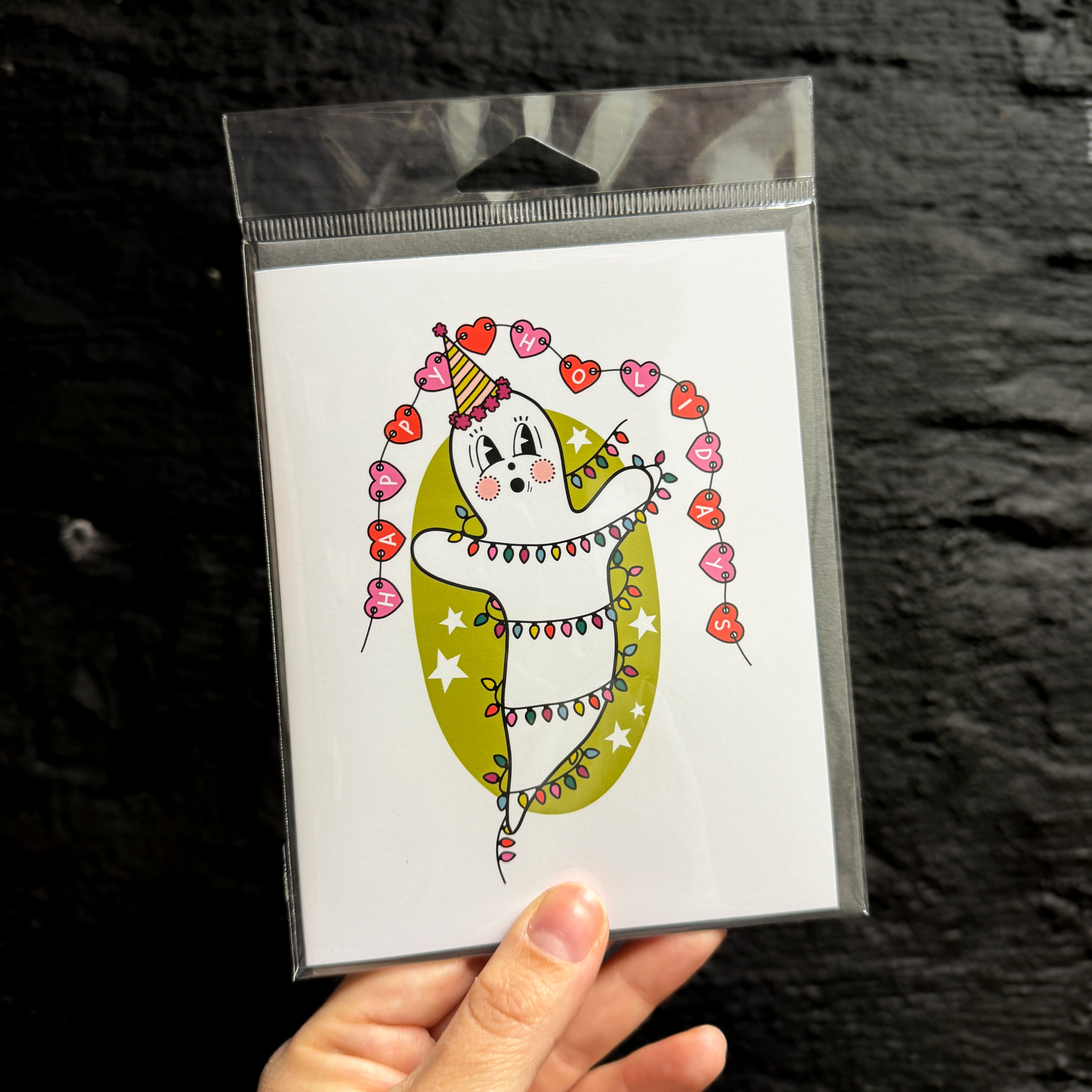 A hand holding a greeting card with a vintage ghost in the middle wearing a party hat, wrapped in Christmas lights, in front of a green oval with white stars. There is a red and pink heart banner above that says 'HAPPY HOLIDAYS'.
