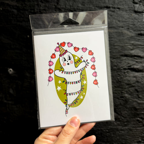 A hand holding a greeting card with a vintage ghost in the middle wearing a party hat, wrapped in Christmas lights, in front of a green oval with white stars. There is a red and pink heart banner above that says 'HAPPY HOLIDAYS'.
