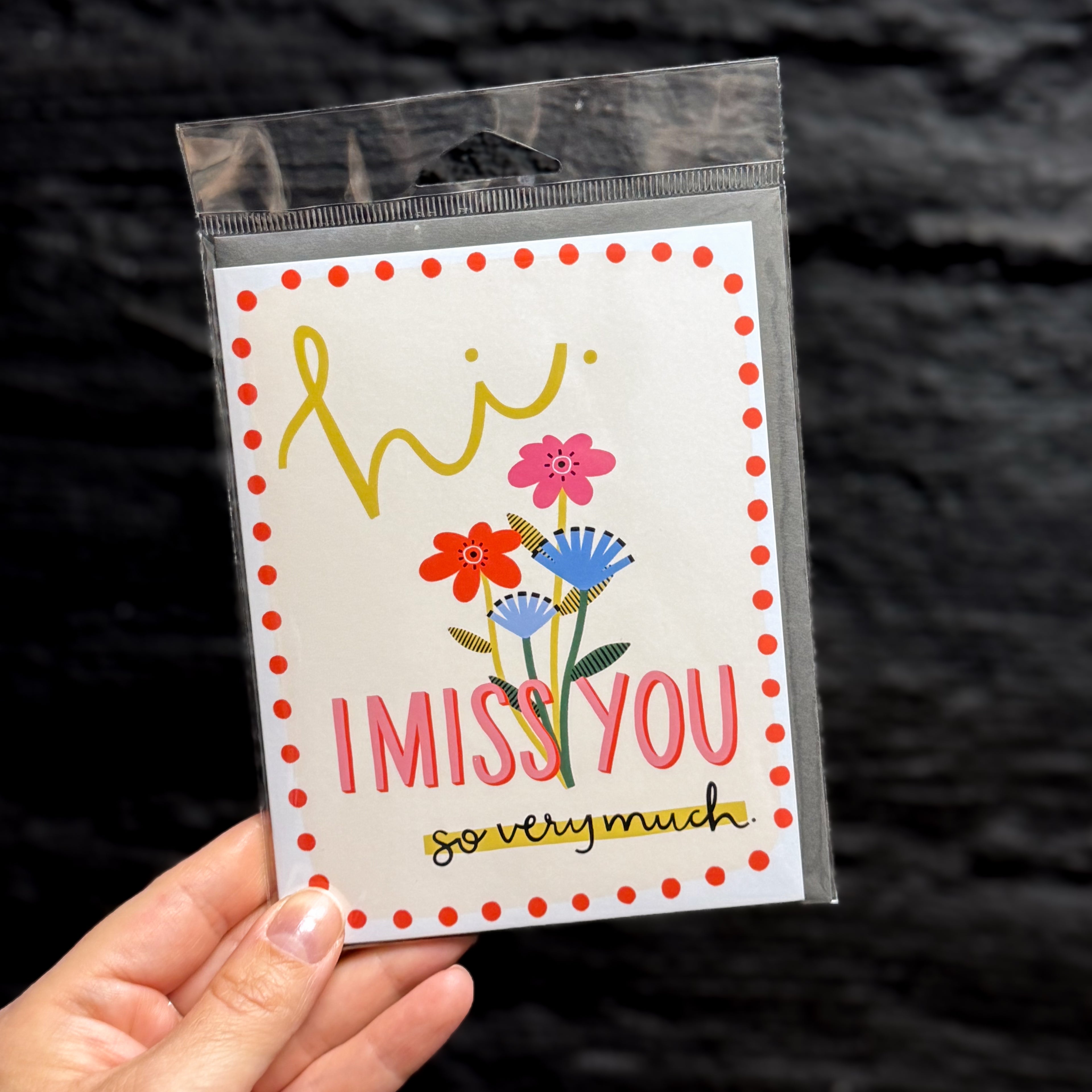A hand holding a packaged greeting card that says 'hi. I MISS YOU so very much.' on a cream background with a red dot border. There are pink and blue flowers in the middle.