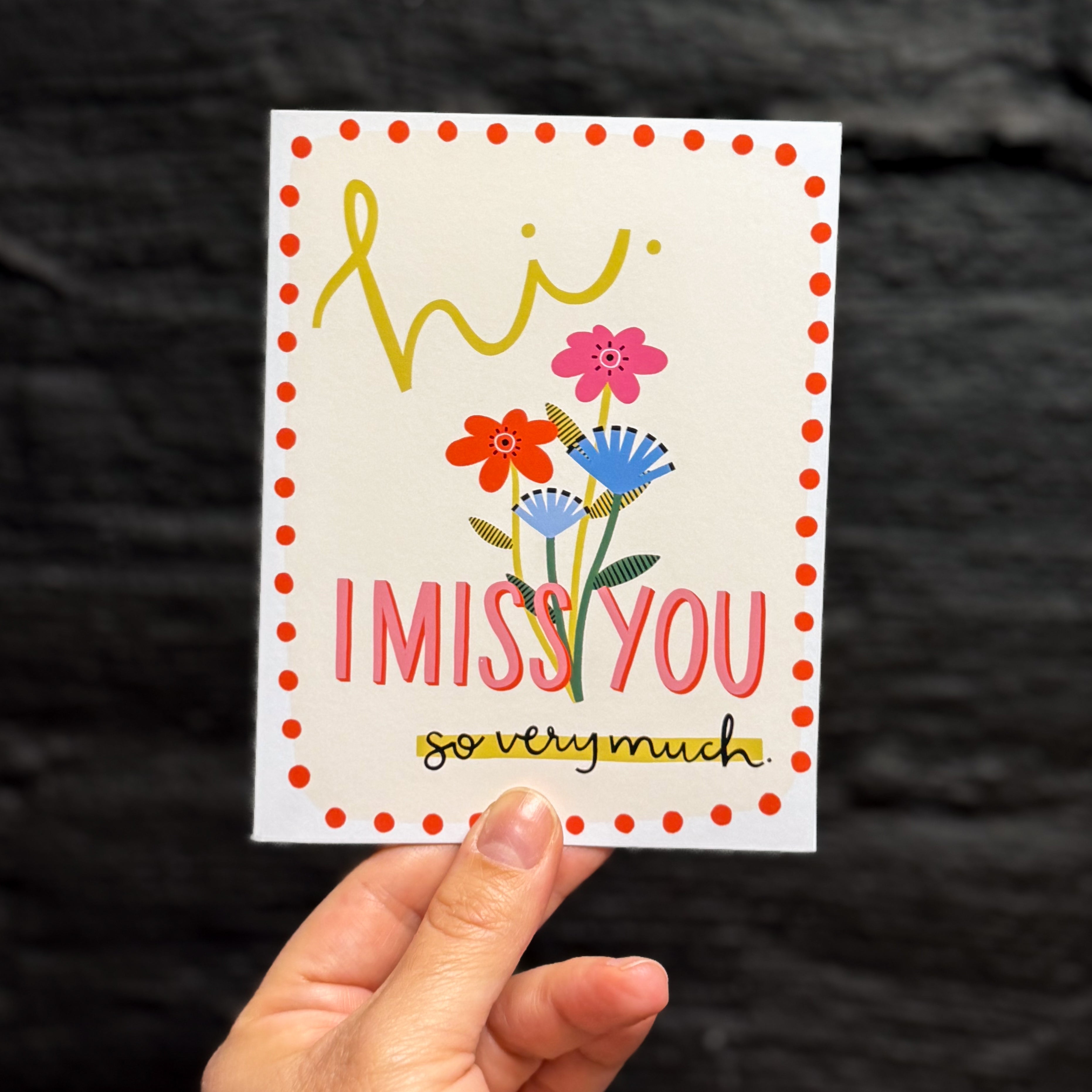 A hand holding a greeting card that says 'hi. I MISS YOU so very much.' on a cream background with a red dot border. There are pink and blue flowers in the middle.