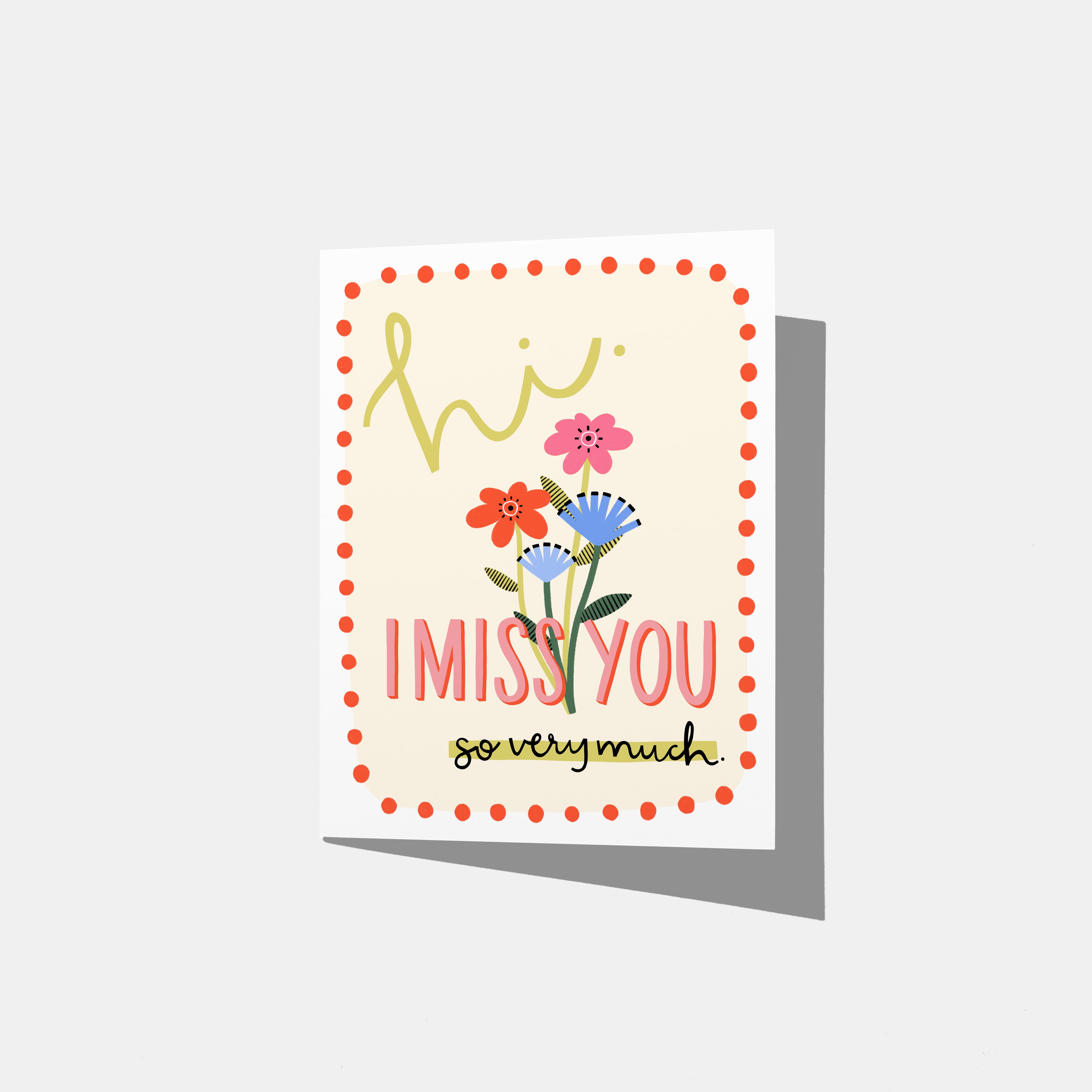 A mockup of a greeting card that says 'hi. I MISS YOU so very much.' on a cream background with a red dot border. There are pink and blue flowers in the middle.