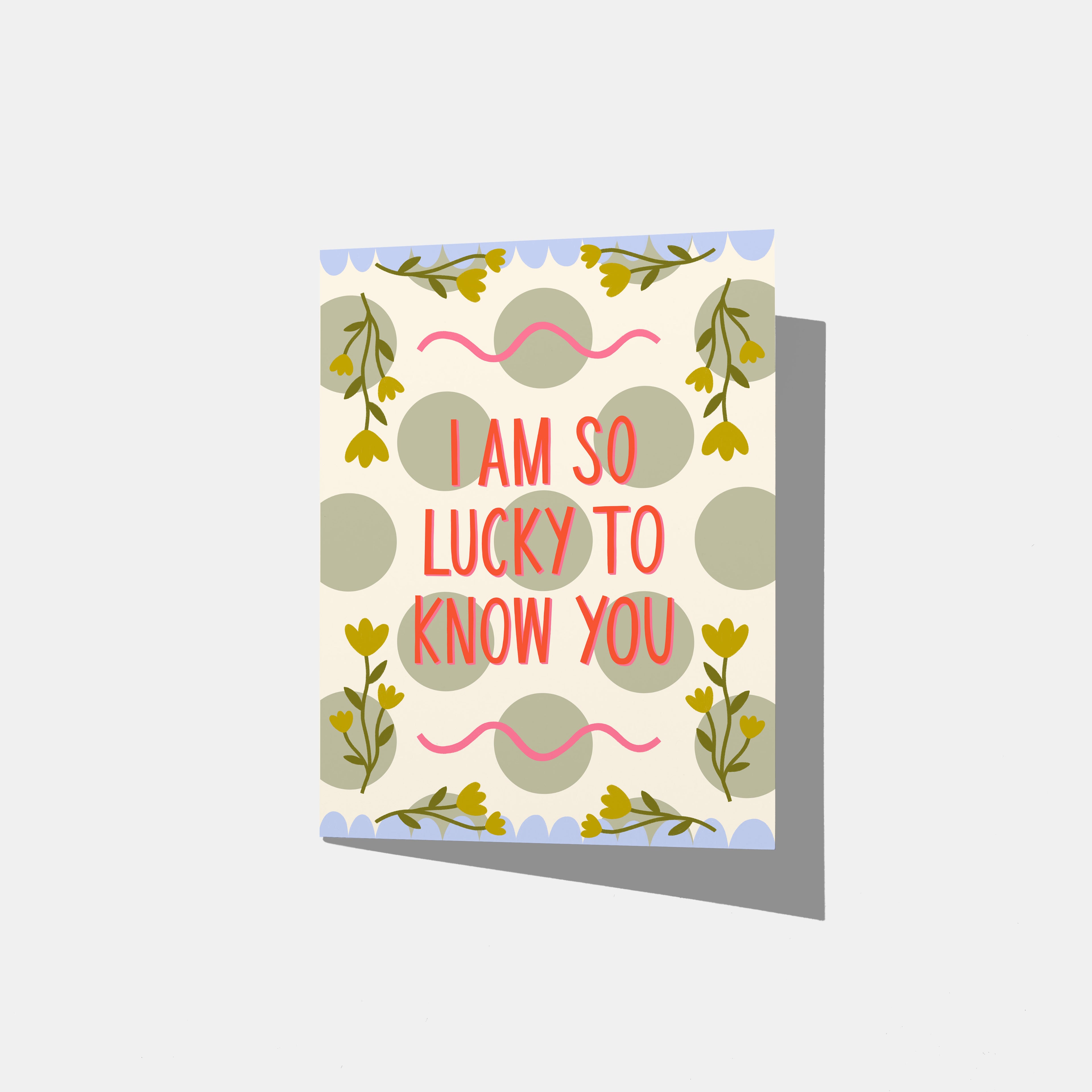 A mockup of a greeting card against a dark textured background. The card has a white base with sage green polka dots, yellow floral sprigs, pink squiggly lines, and red text that reads, 'I AM SO LUCKY TO KNOW YOU'.
