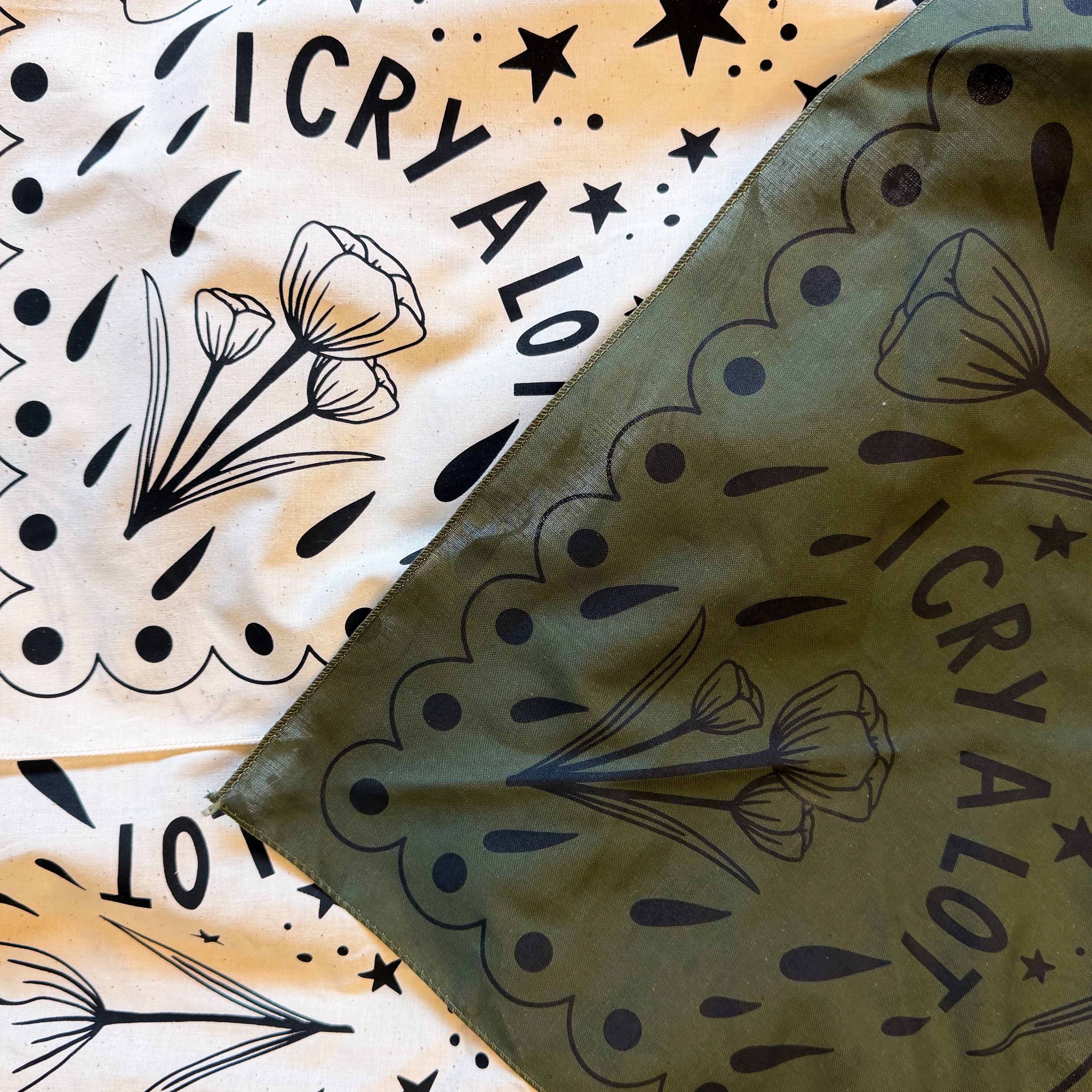 One Natural and one olive color bandanas with black floral patterns and 'I CRY A LOT' text.