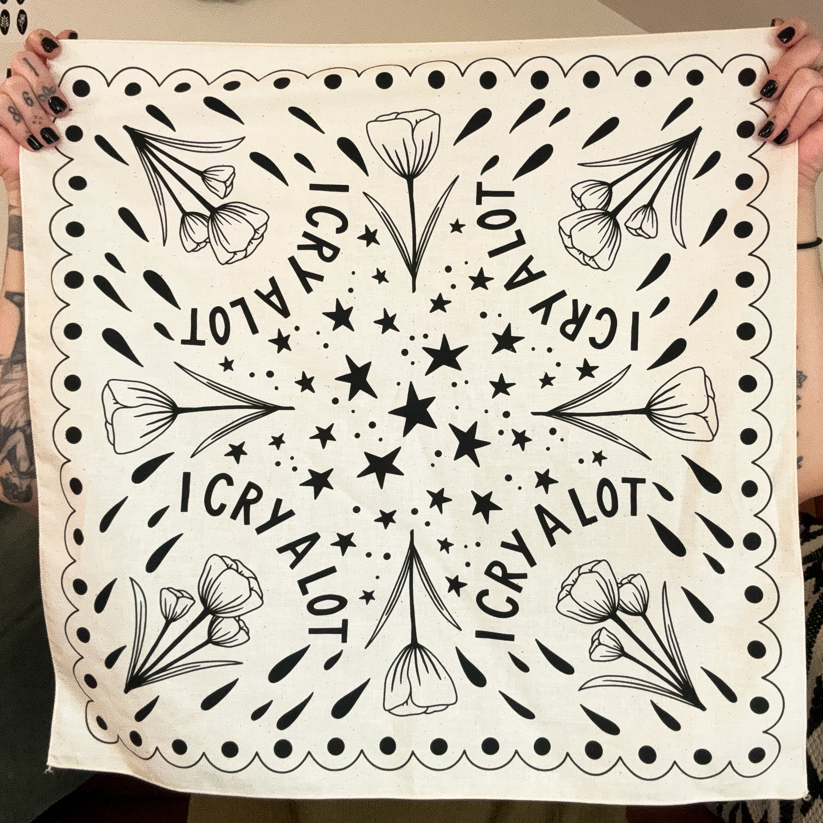 Natural color bandana with black floral patterns and 'I CRY A LOT' text held up by two hands.