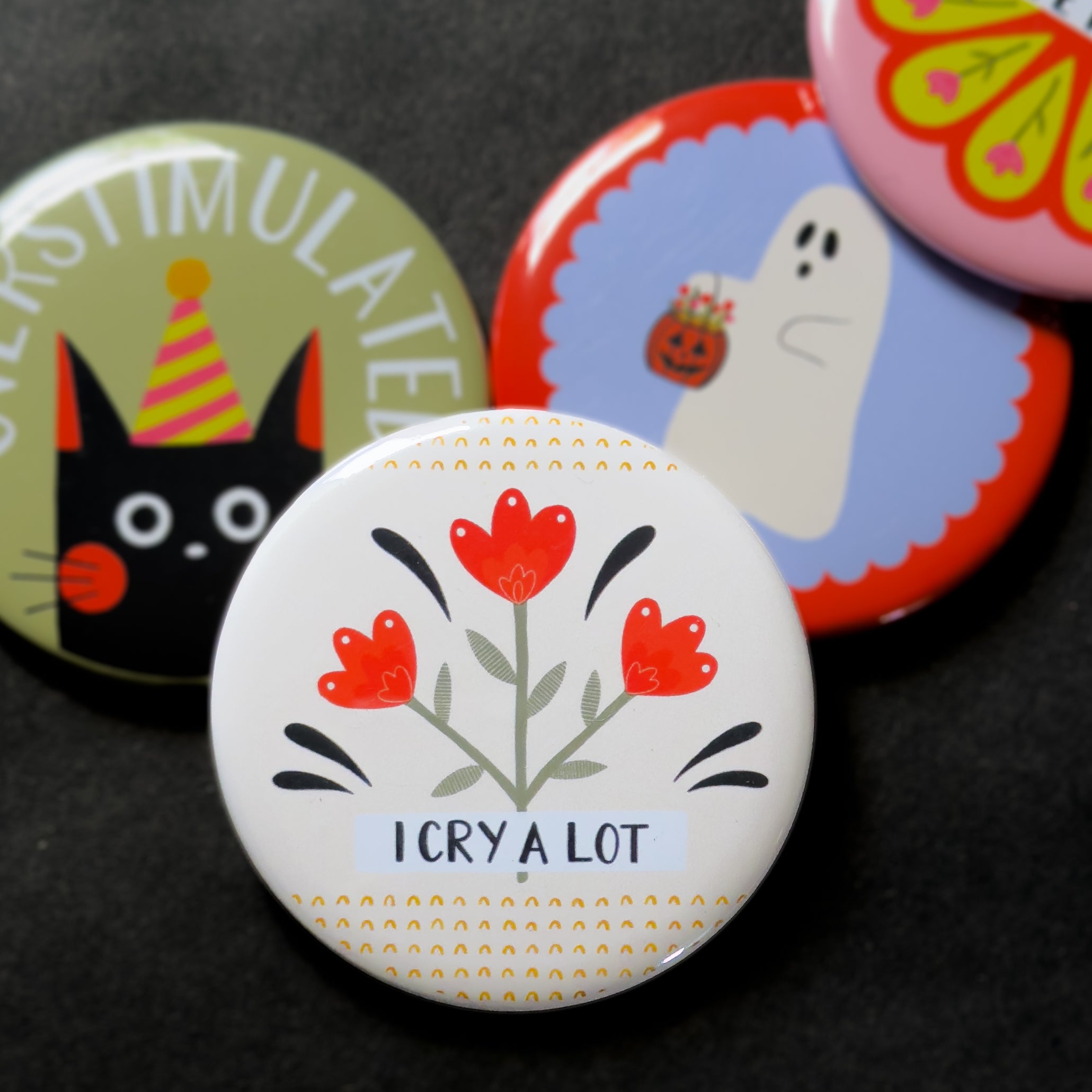 A round pinback button featuring a cream background with red flowers and black and yellow embellishments and the text 'I CRY A LOT'. The pin is surrounded by other pin designs.