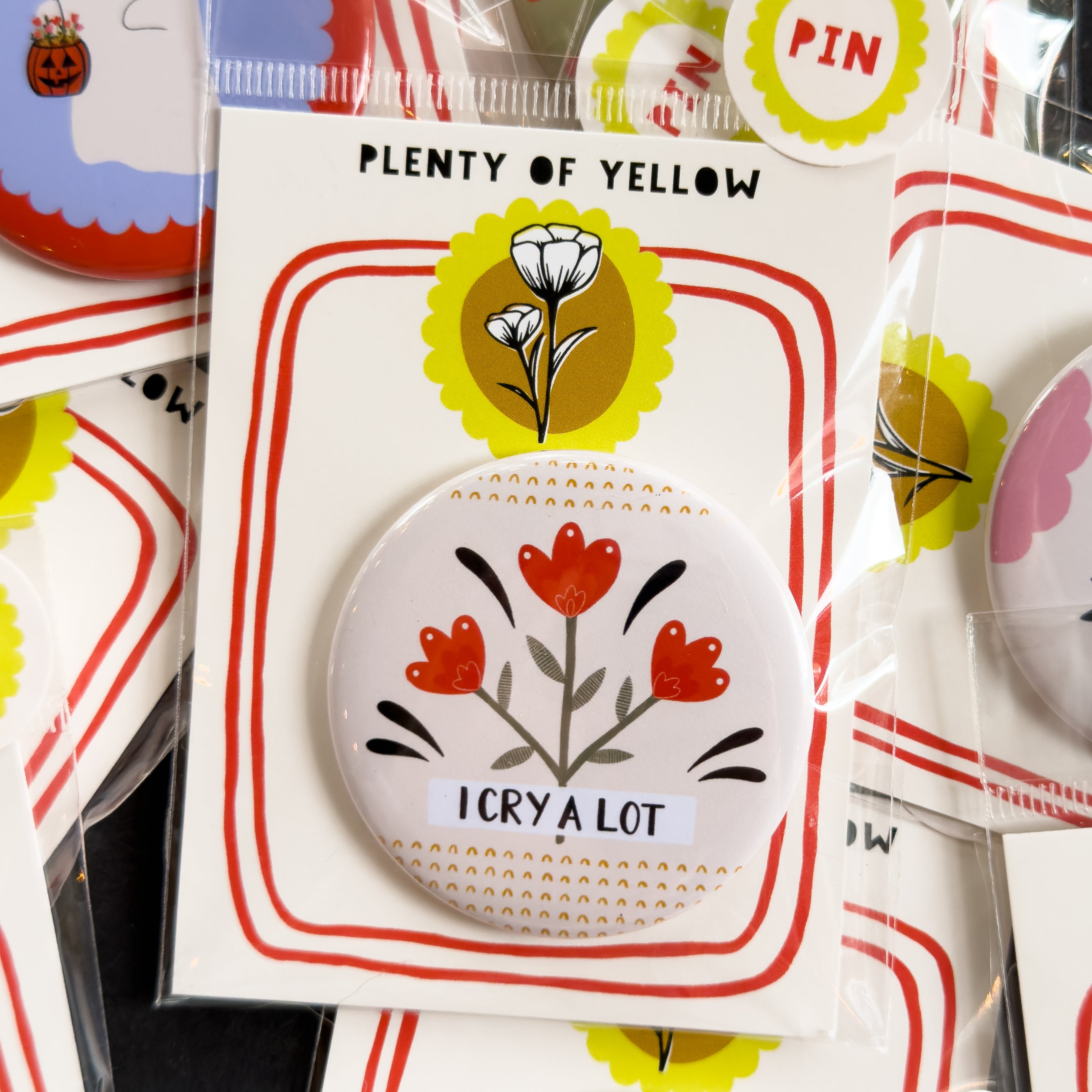 A round pinback button in clear plastic packaging, featuring a cream background with red flowers and black and yellow embellishments and the text 'I CRY A LOT'. The pin is mounted on a white backing card with red borders and a yellow logo, under the brand name 'PLENTY OF YELLOW'.