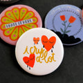A round pinback button featuring a cream background with red crying hearts and the text ‘i cry a lot'. The pin is surrounded by other pin designs.