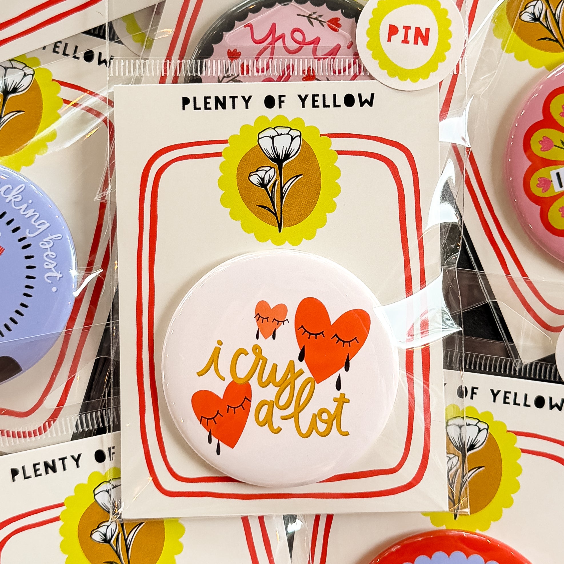 A round pinback button in clear plastic packaging, featuring a cream background with red crying hearts and the text ‘i cry a lot’. The pin is mounted on a white backing card with red borders and a yellow logo, under the brand name 'PLENTY OF YELLOW'.