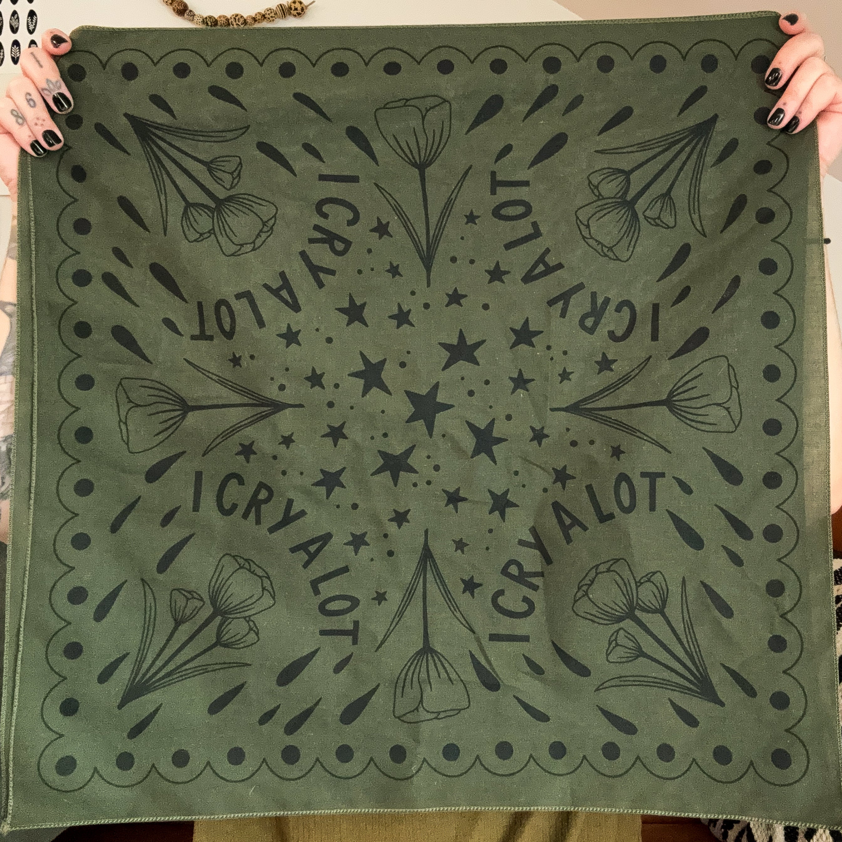 Olive green bandana with black floral patterns and 'I CRY A LOT' text held up by two hands.