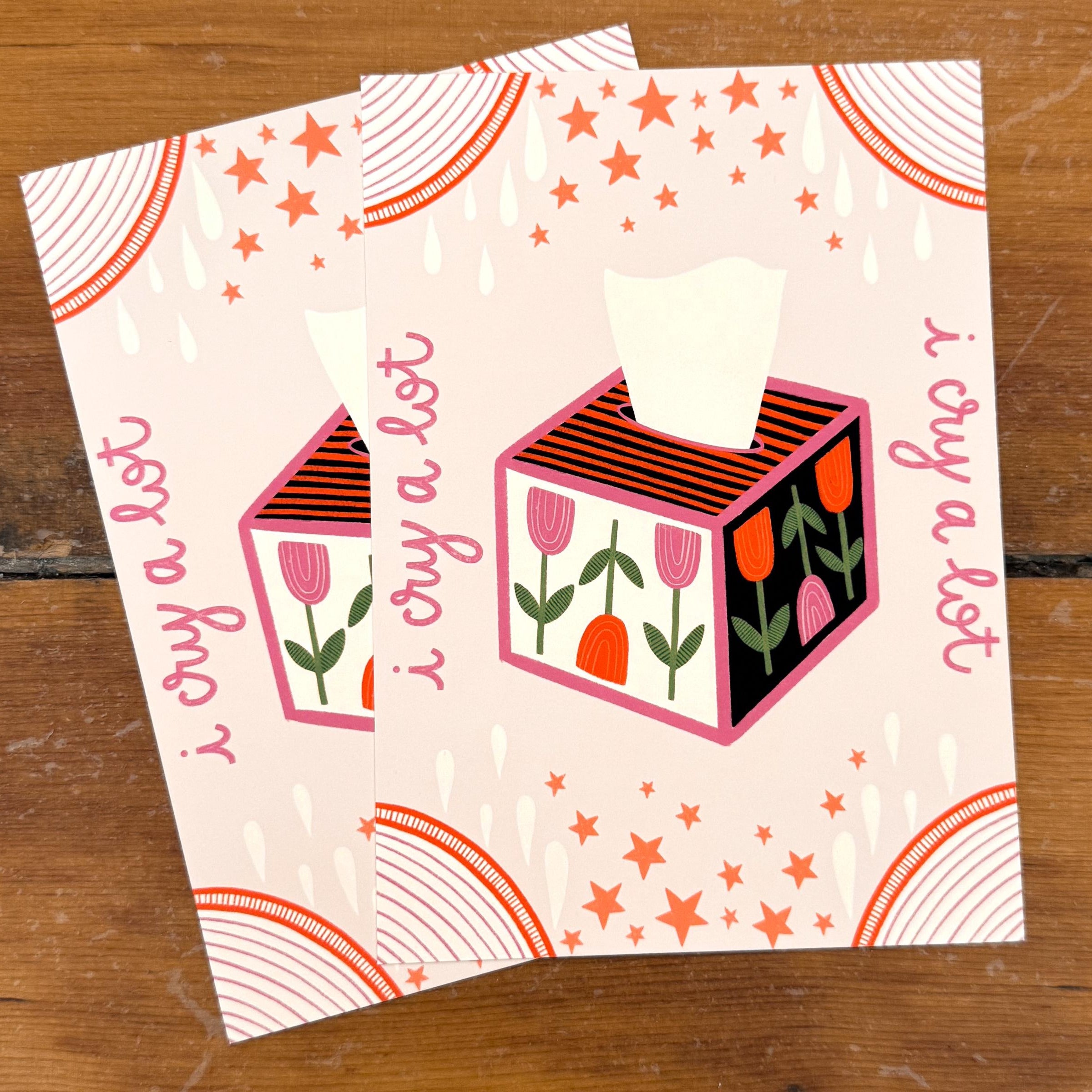 Two 5x7 art prints that say 'i cry a lot' on each side with a floral tissue box in the middle. Embellished with a pink background, peach details, and tear drops. Sitting on a wood background.