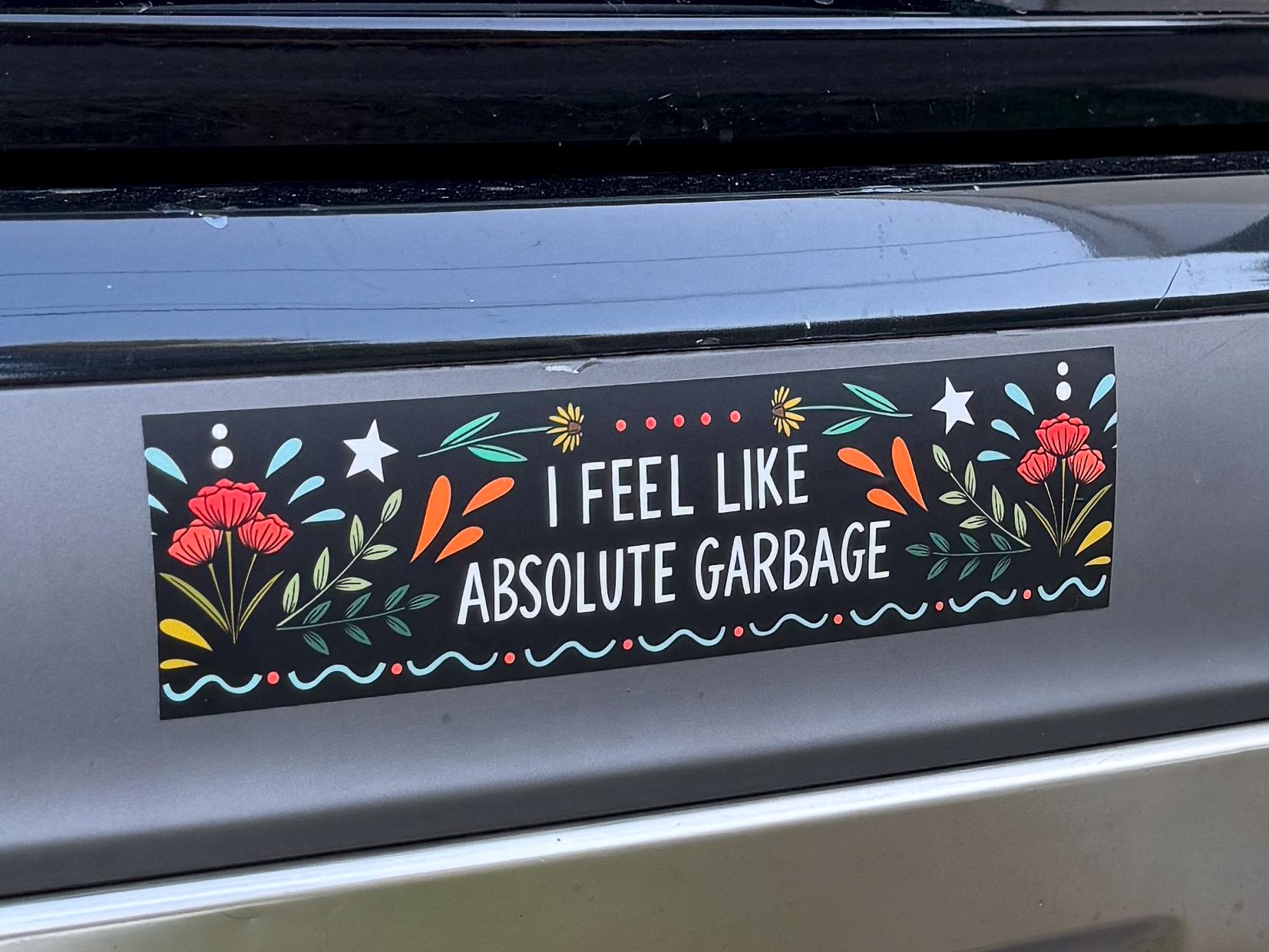 A black bumper sticker that says 'I FEEL LIKE ABSOLUTE GARBAGE' surrounded by flowers, starts, and whimsy designs on a car bumper.