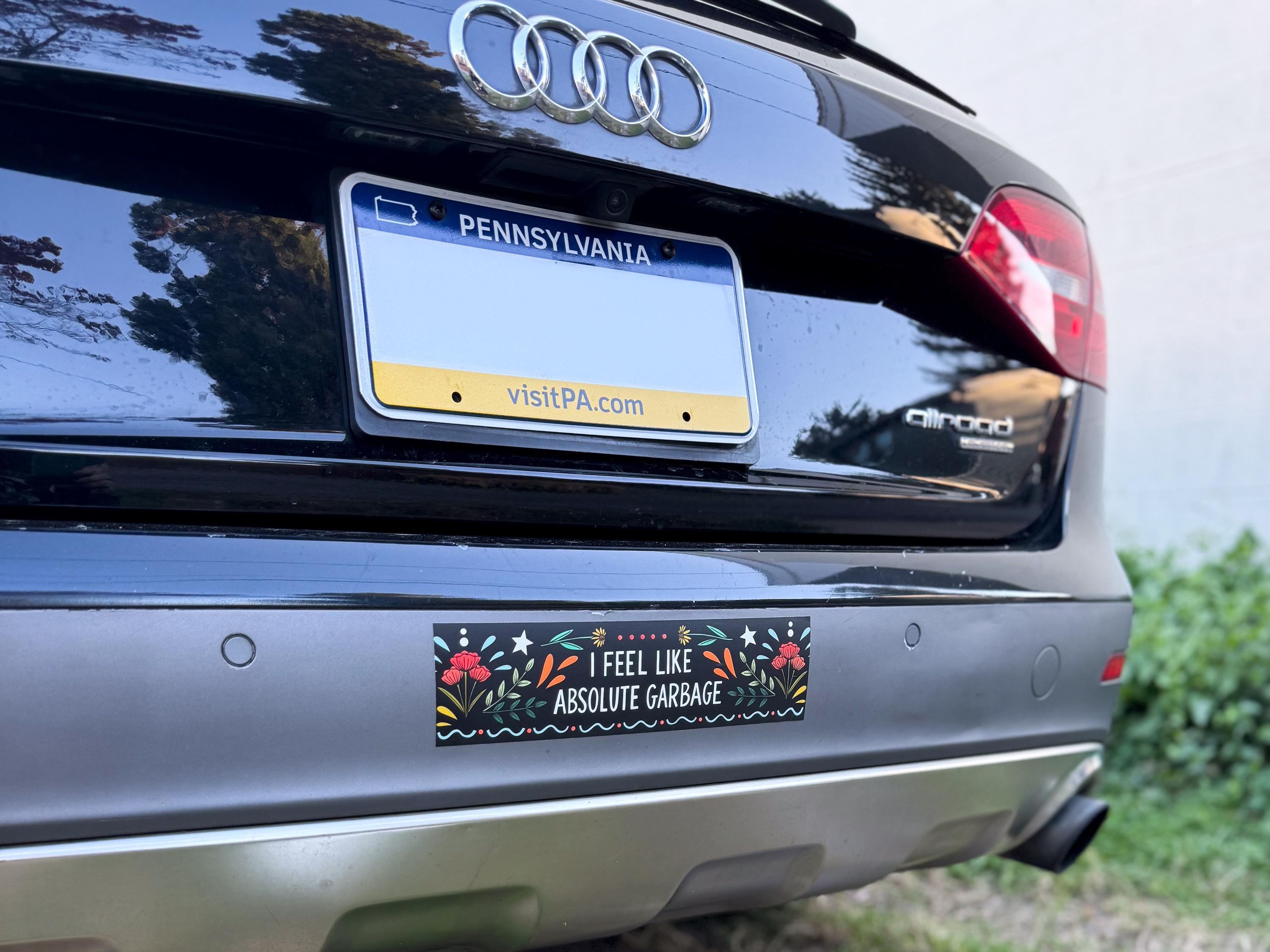 The back of a black Audi allroad that has a black bumper sticker in the middle of the bumper that says 'I FEEL LIKE ABSOLUTE GARBAGE' surrounded by flowers, starts, and whimsy designs.