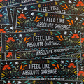 A black bumper sticker that says 'I FEEL LIKE ABSOLUTE GARBAGE' surrounded by flowers, starts, and whimsy designs.