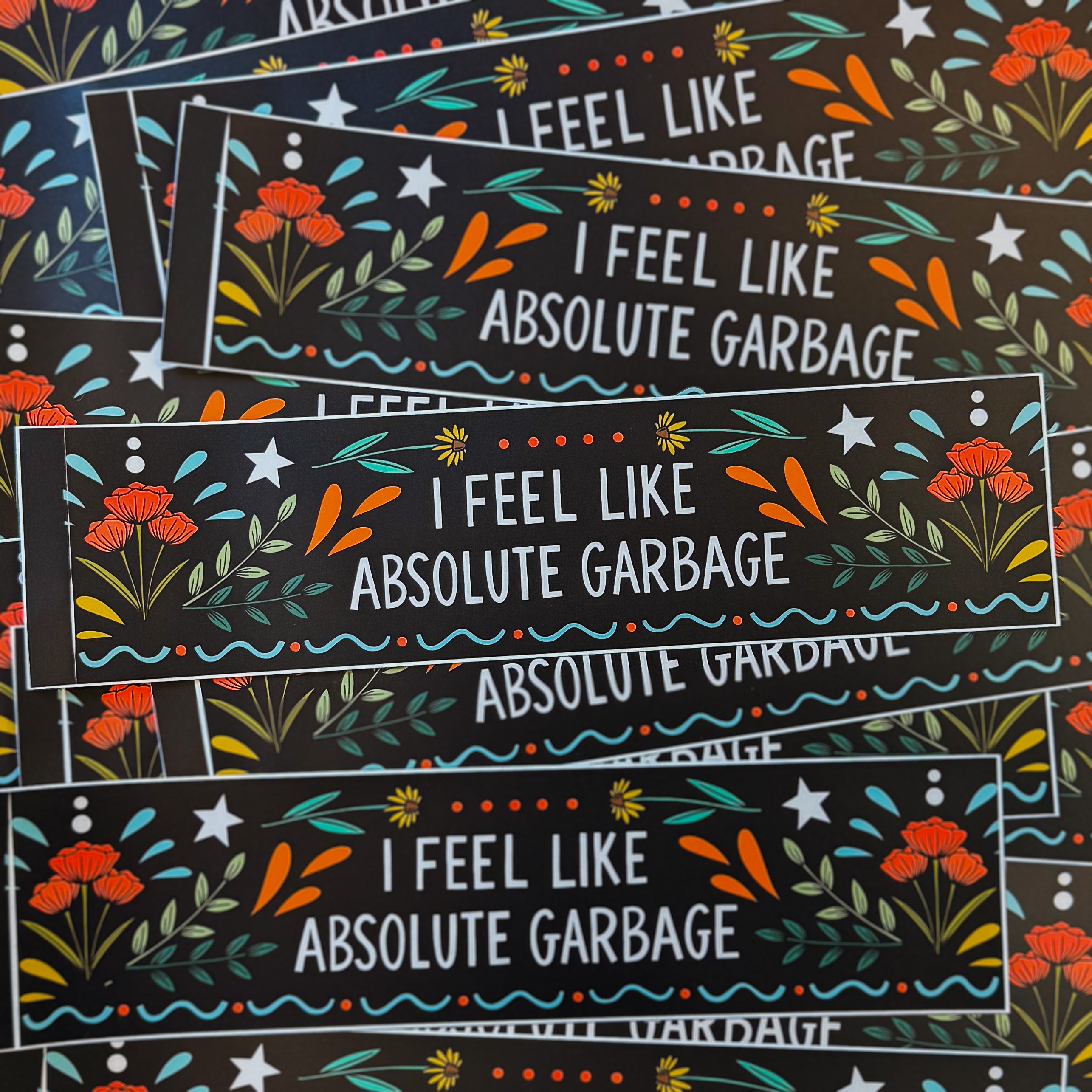 A black bumper sticker that says 'I FEEL LIKE ABSOLUTE GARBAGE' surrounded by flowers, starts, and whimsy designs.