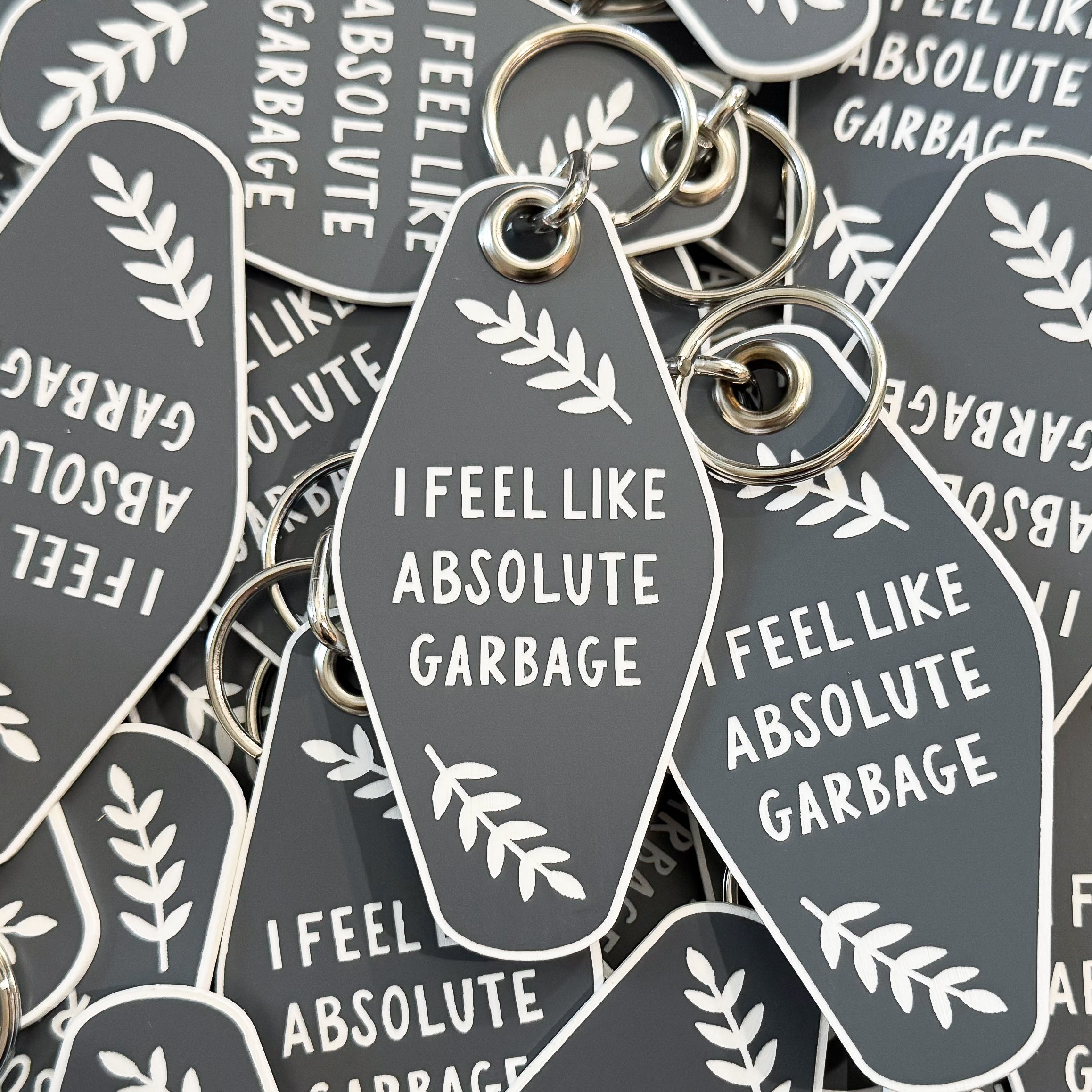 A pile of motel-style keychains in grey with a white border that say 'I FEEL LIKE ABSOLUTE GARBAGE' in the middle surrounded by leaves.